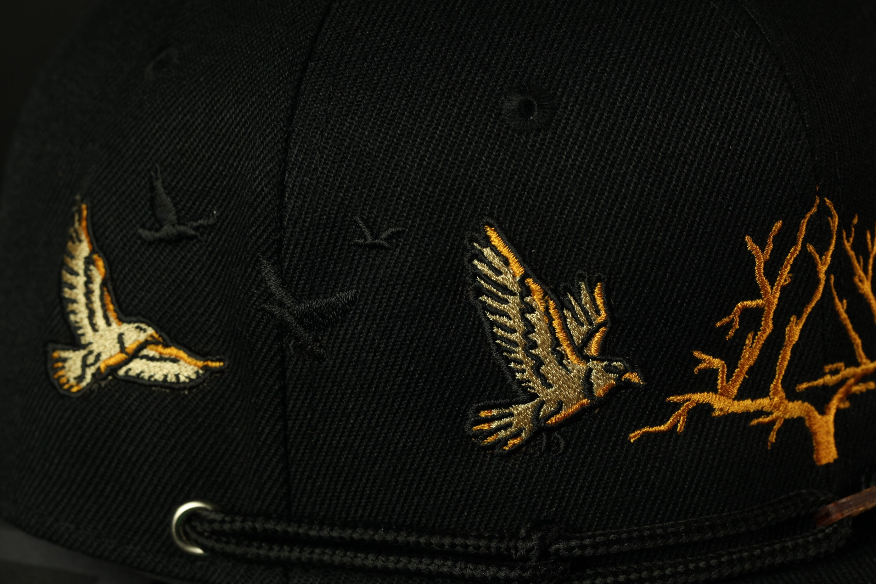 a close-up view of a black baseball cap with gold embroidered designs. The designs include a bird in flight, a bird perched on a branch, and a bird in flight.