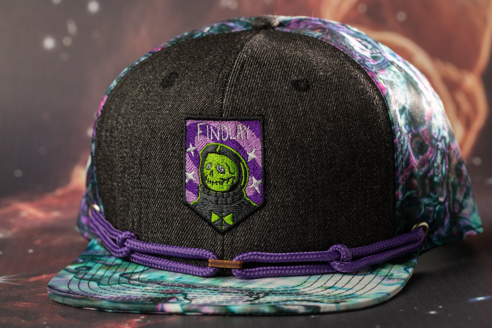 A black baseball cap with a purple and green patterned design, featuring a skull and crossbones motif.
