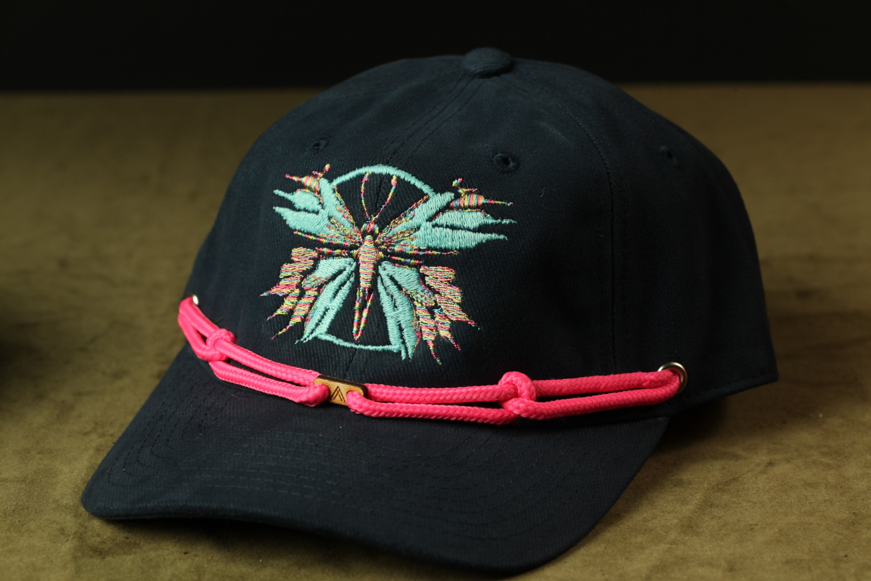A black baseball cap with a pink braided strap and a colorful embroidered design of a butterfly on the front.
