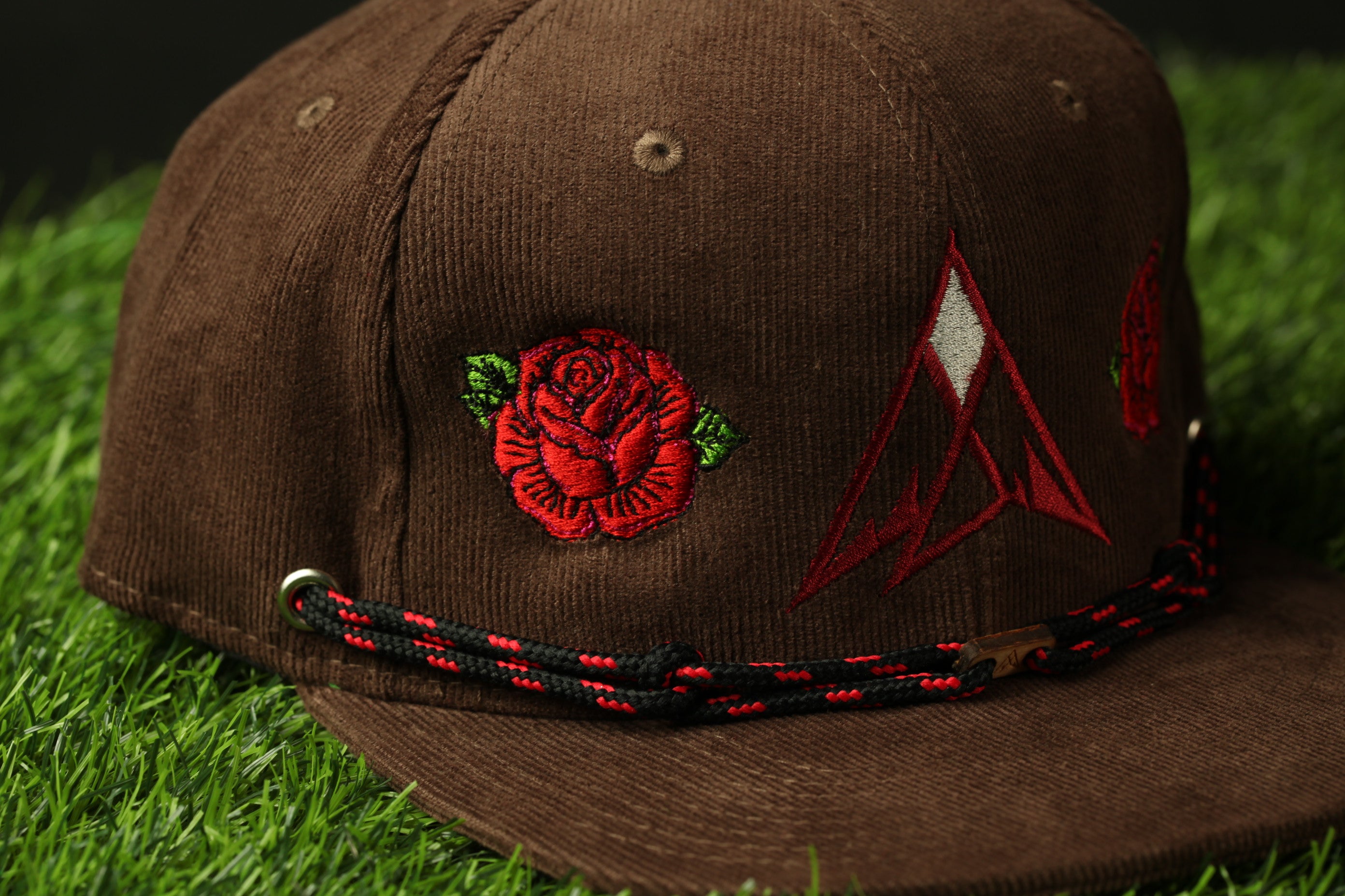 A brown baseball cap with a red rose embroidered on the front and a red triangle design on the brim, sitting on a grassy surface.