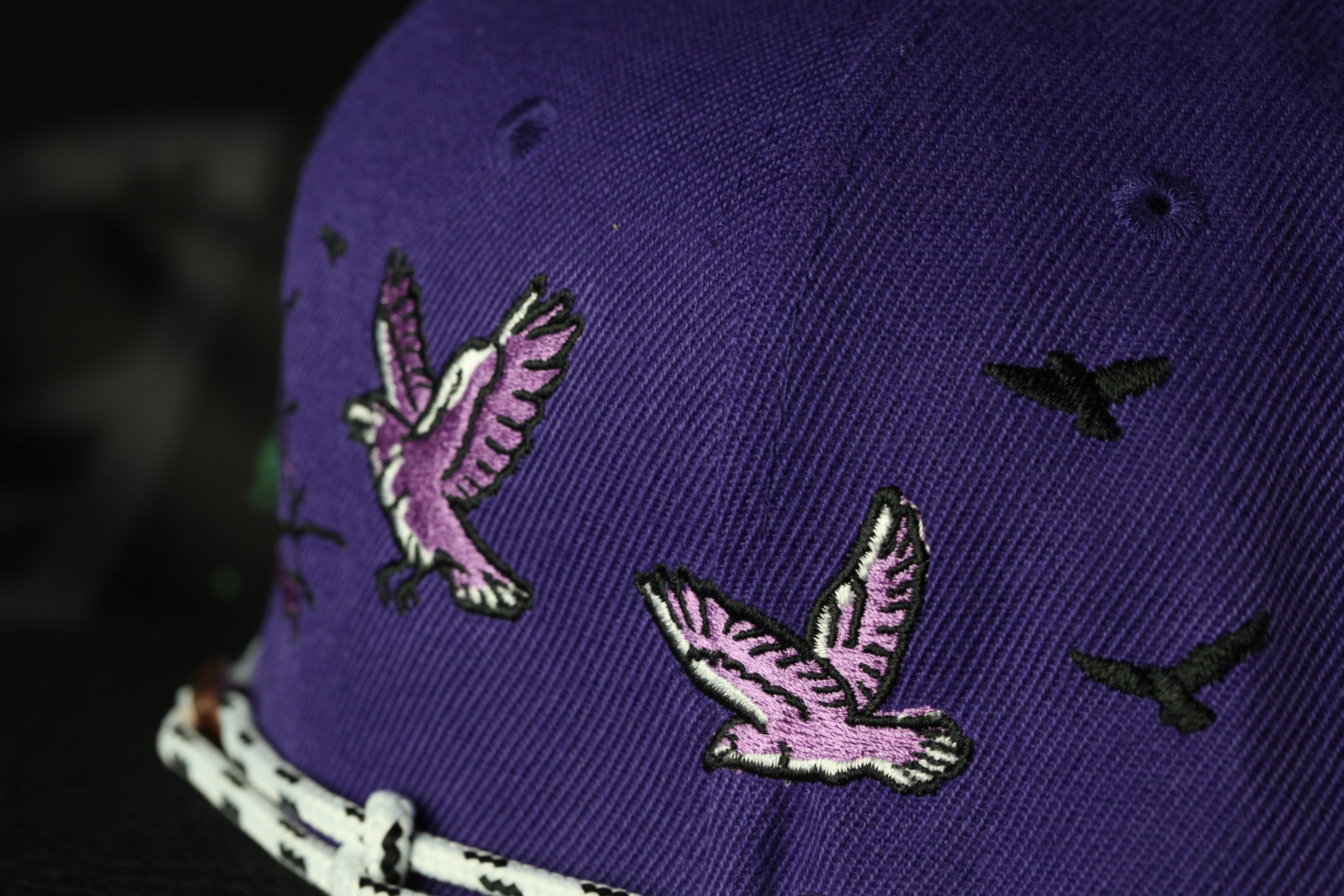 A close-up view of a purple baseball cap with a white border, featuring embroidered images of purple birds in flight.