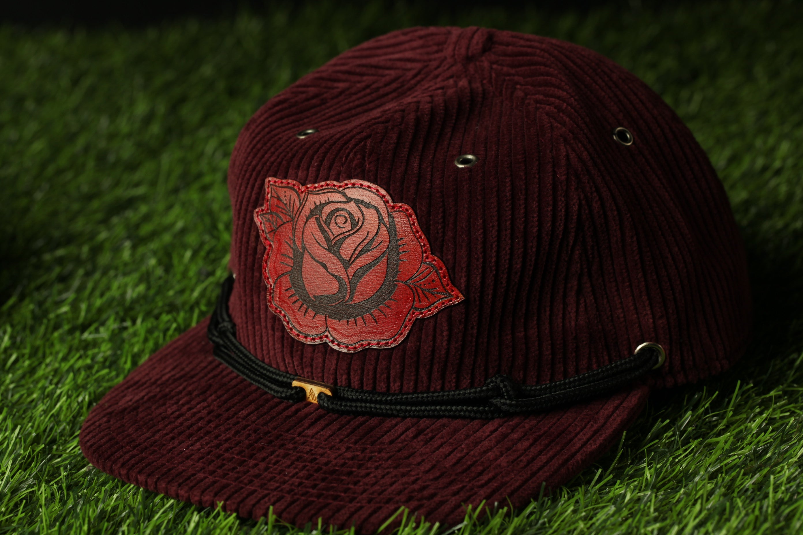 A burgundy baseball cap with a red rose embroidered on the front, resting on a grassy surface.