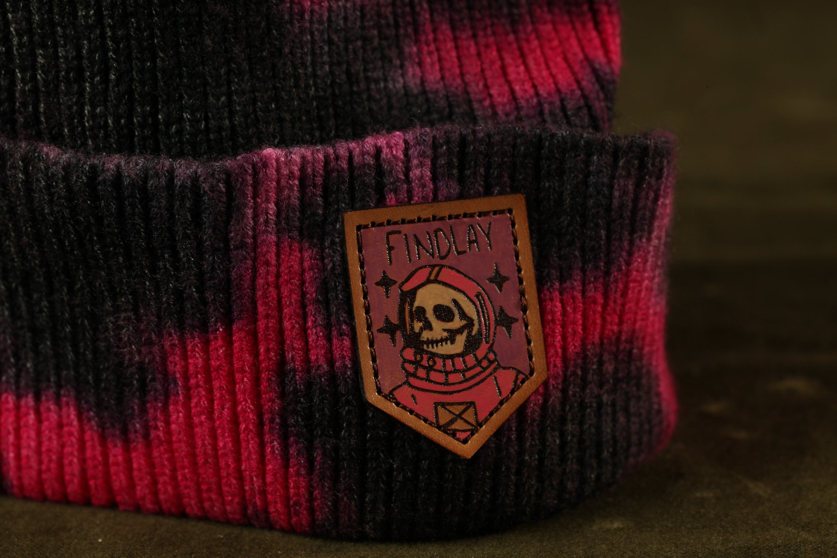 A close-up of a knitted or crocheted beanie with a pink and black striped pattern. The beanie has a patch on it that features a skull wearing a red scarf and the word "FINDLAY" written on it.