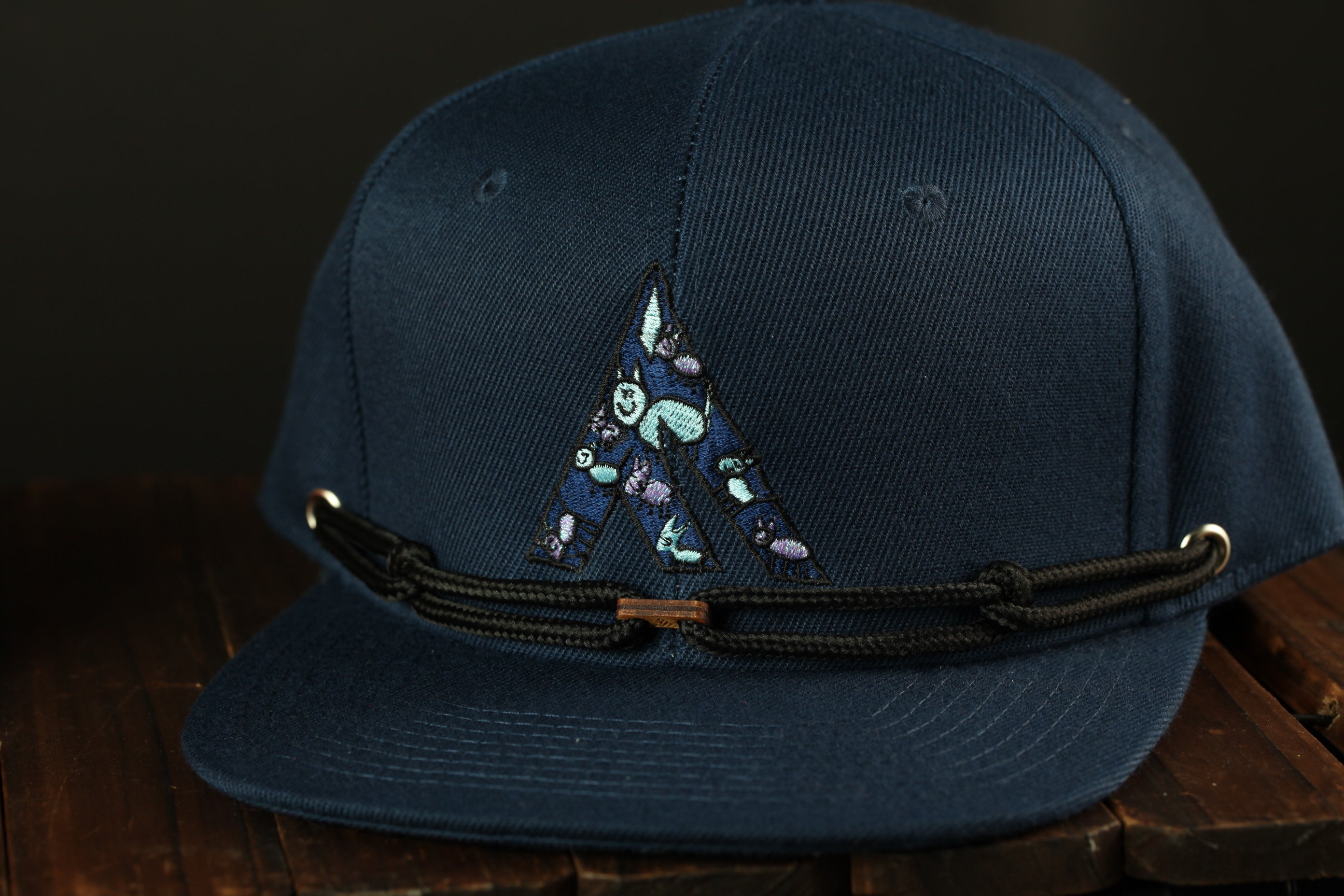 A navy blue baseball cap with a decorative embroidered letter "A" on the front, sitting on a wooden surface.