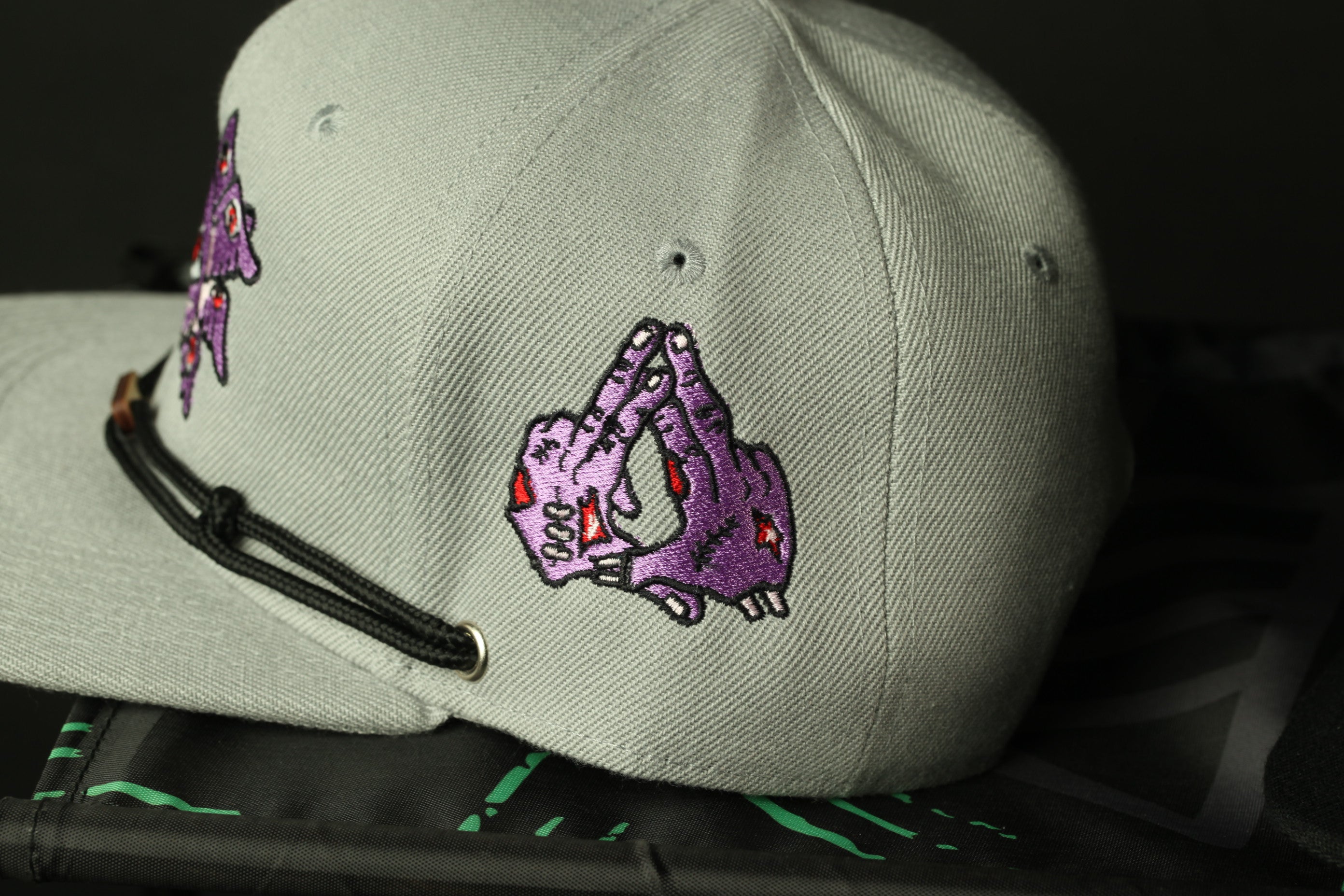 A close-up view of a gray baseball cap with a purple and pink embroidered design on the front.
