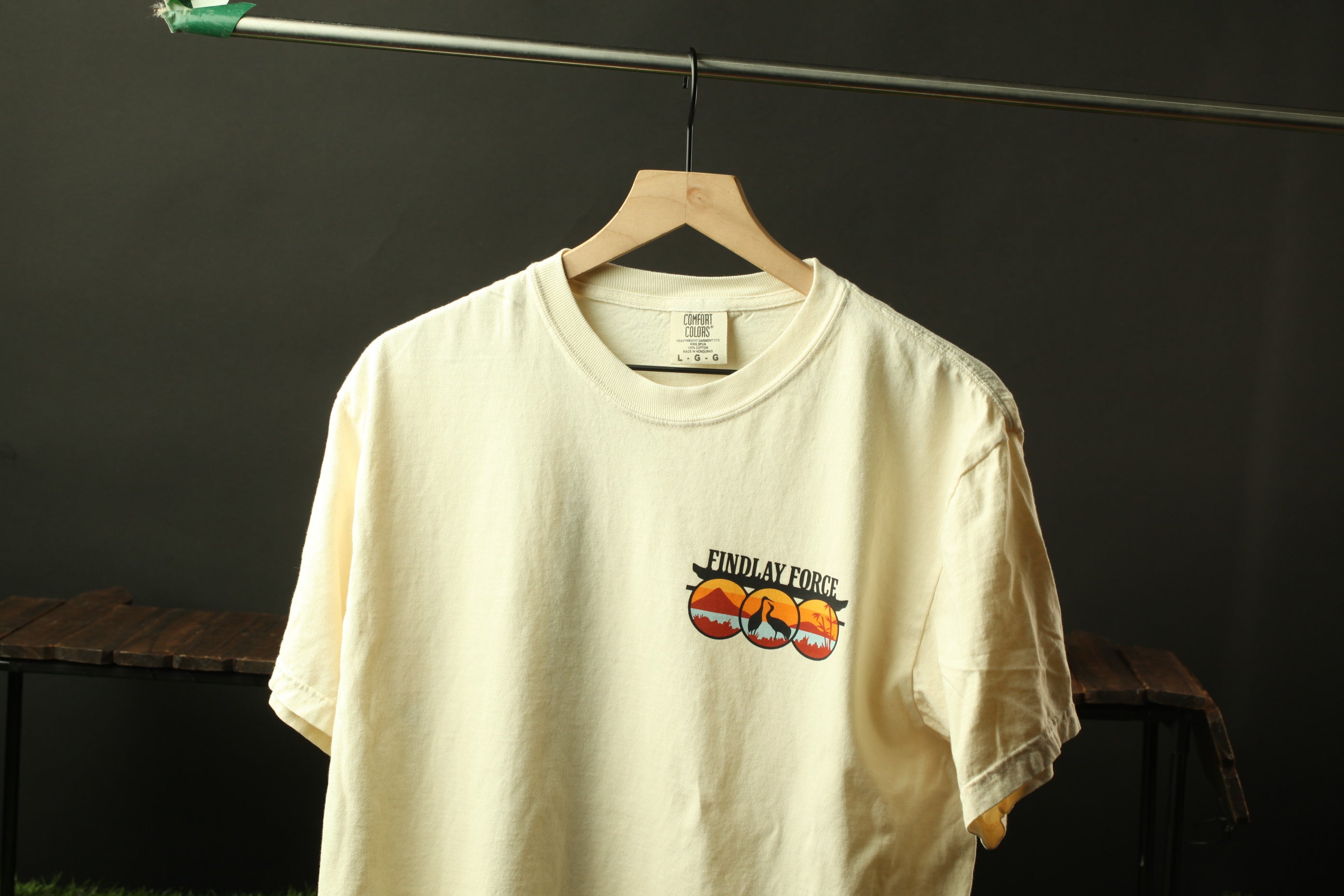 A white t-shirt with a graphic design hanging on a wooden hanger against a dark background.