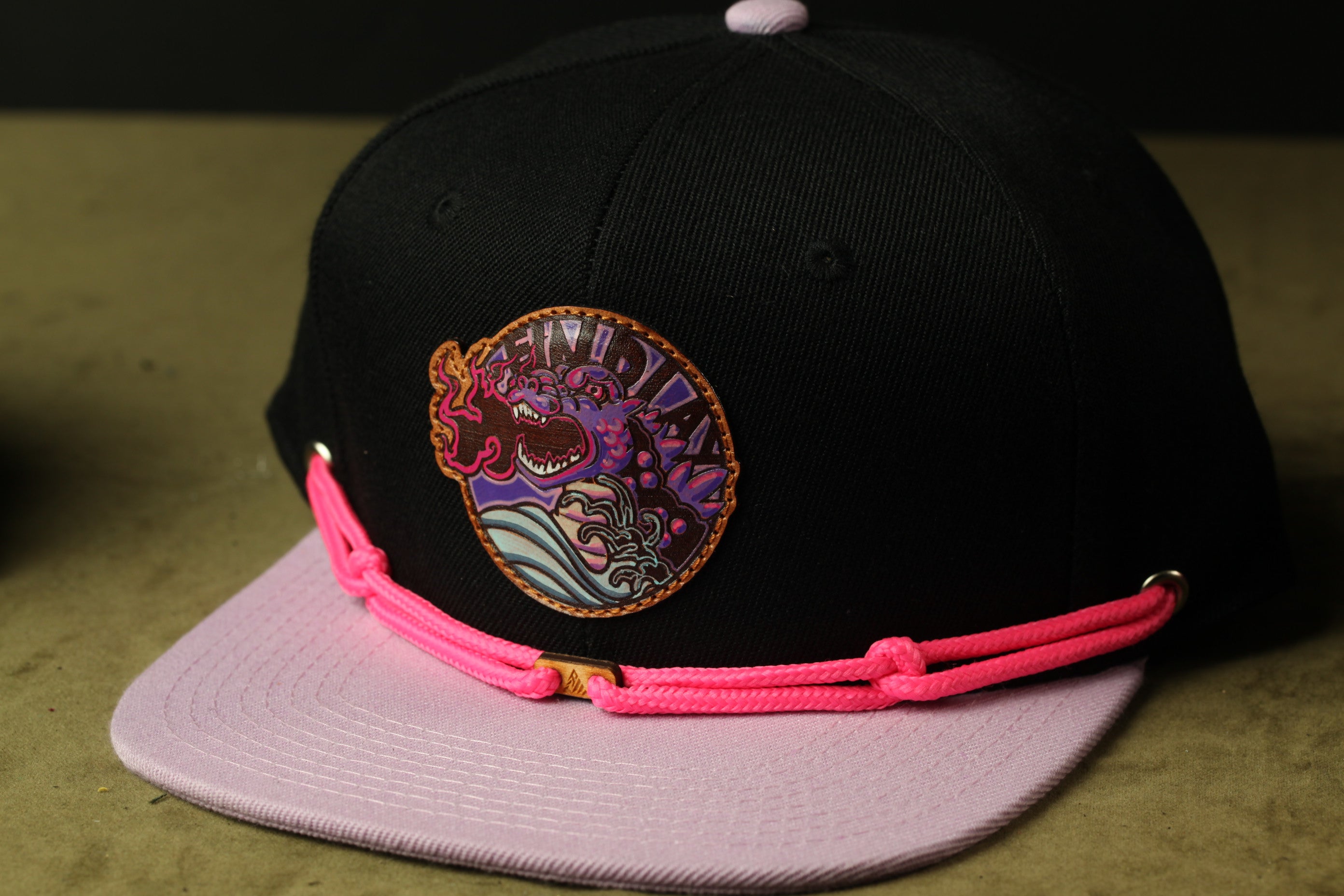 A black baseball cap with a pink and purple design on the front, featuring a circular emblem with a dragon and waves.