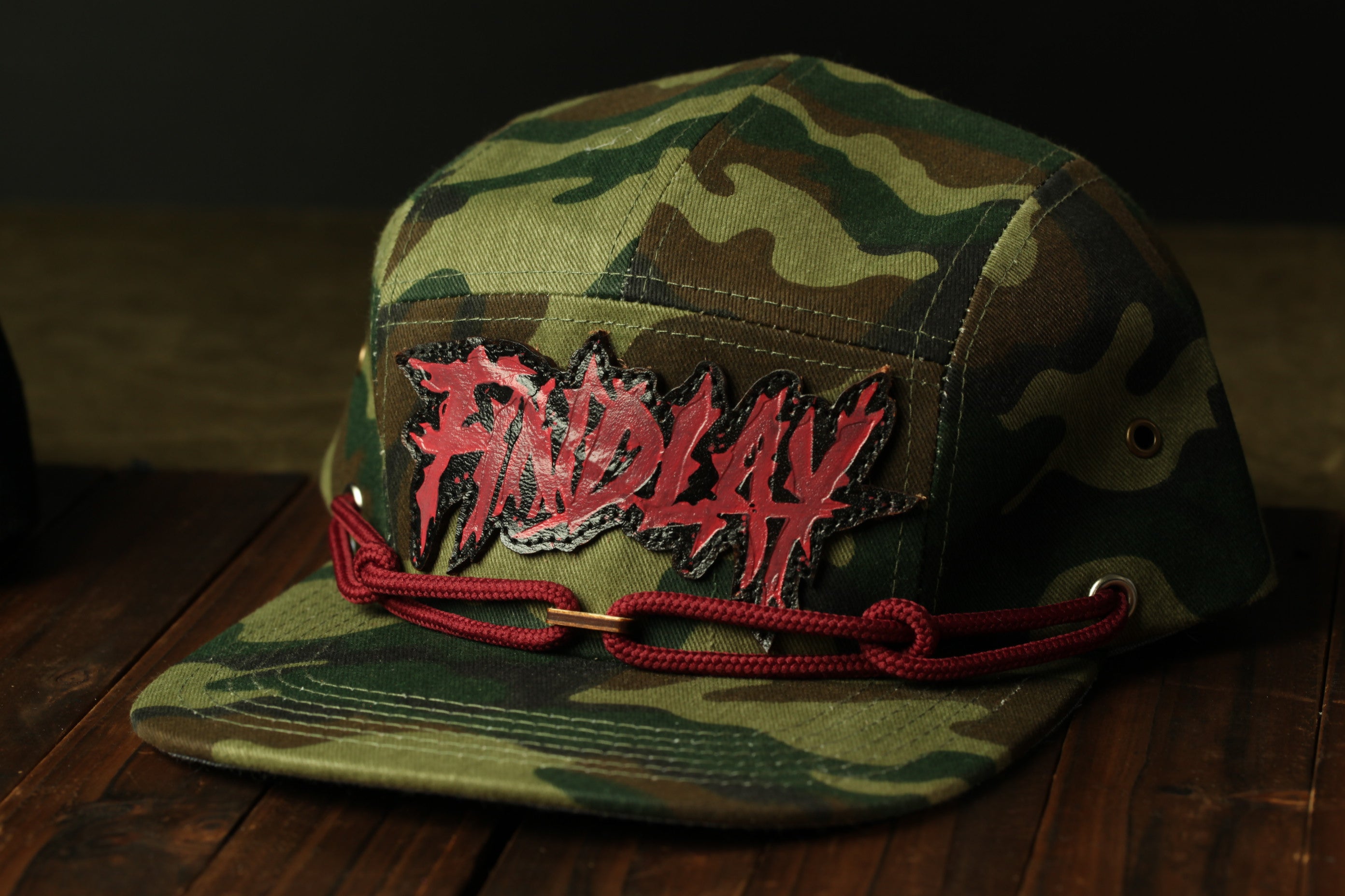 A camouflage baseball cap with a red and black graphic design on the front, sitting on a wooden surface.