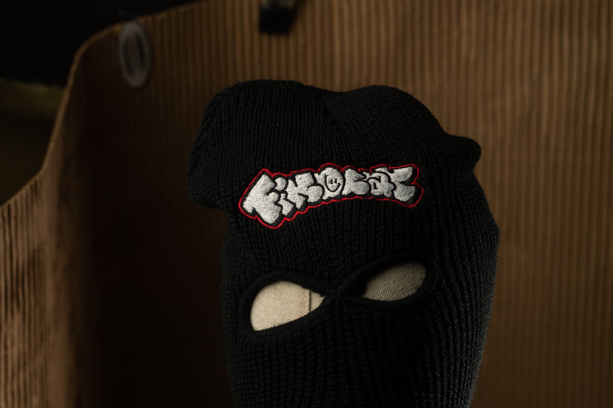A black ski mask with a red and white embroidered logo on the front.