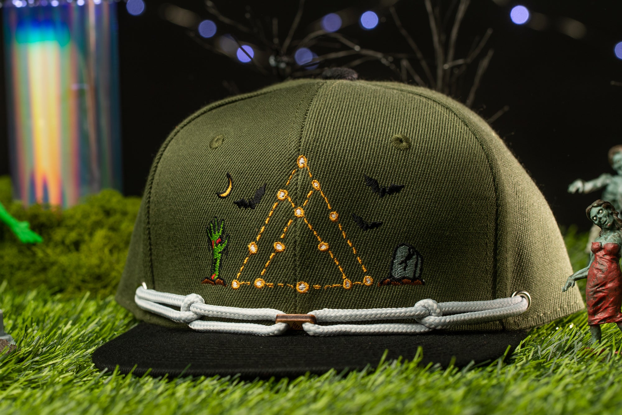 A green baseball cap with a unique design featuring a triangle, a bat, a skull, and a plant, all set against a dark background with a few small figures and a colorful object in the background.