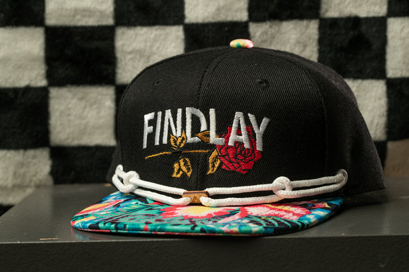 A black baseball cap with a floral pattern and the word "FINDPLAY" embroidered on it, placed on a table against a checkered background.