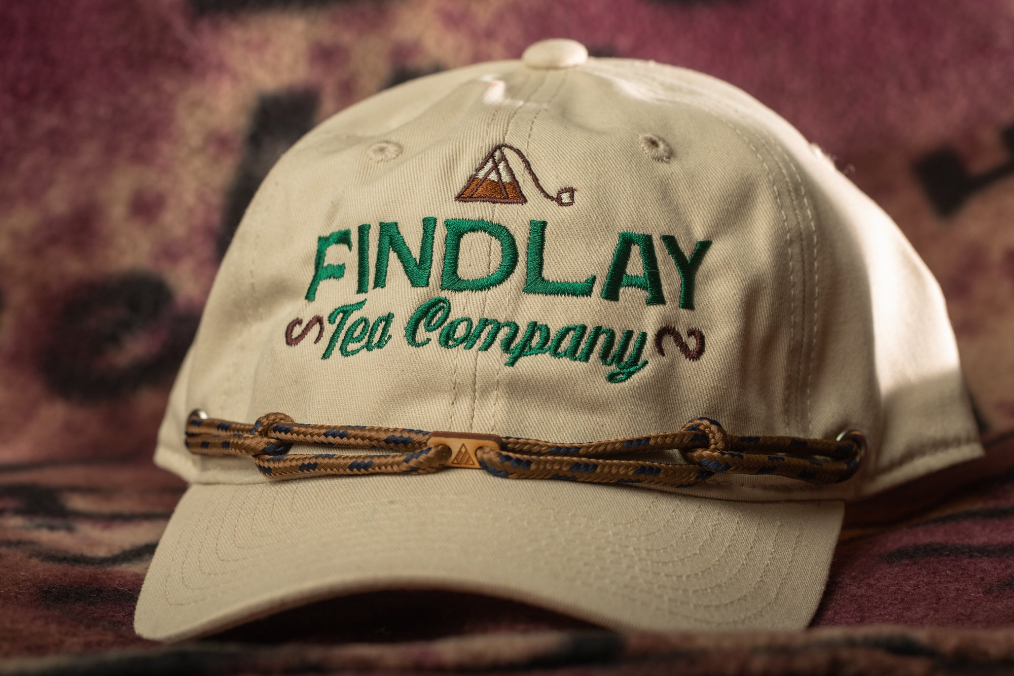 A beige baseball cap with the text "FIND LAY" and "Tea Company" embroidered on it, along with a rope-like strap.
