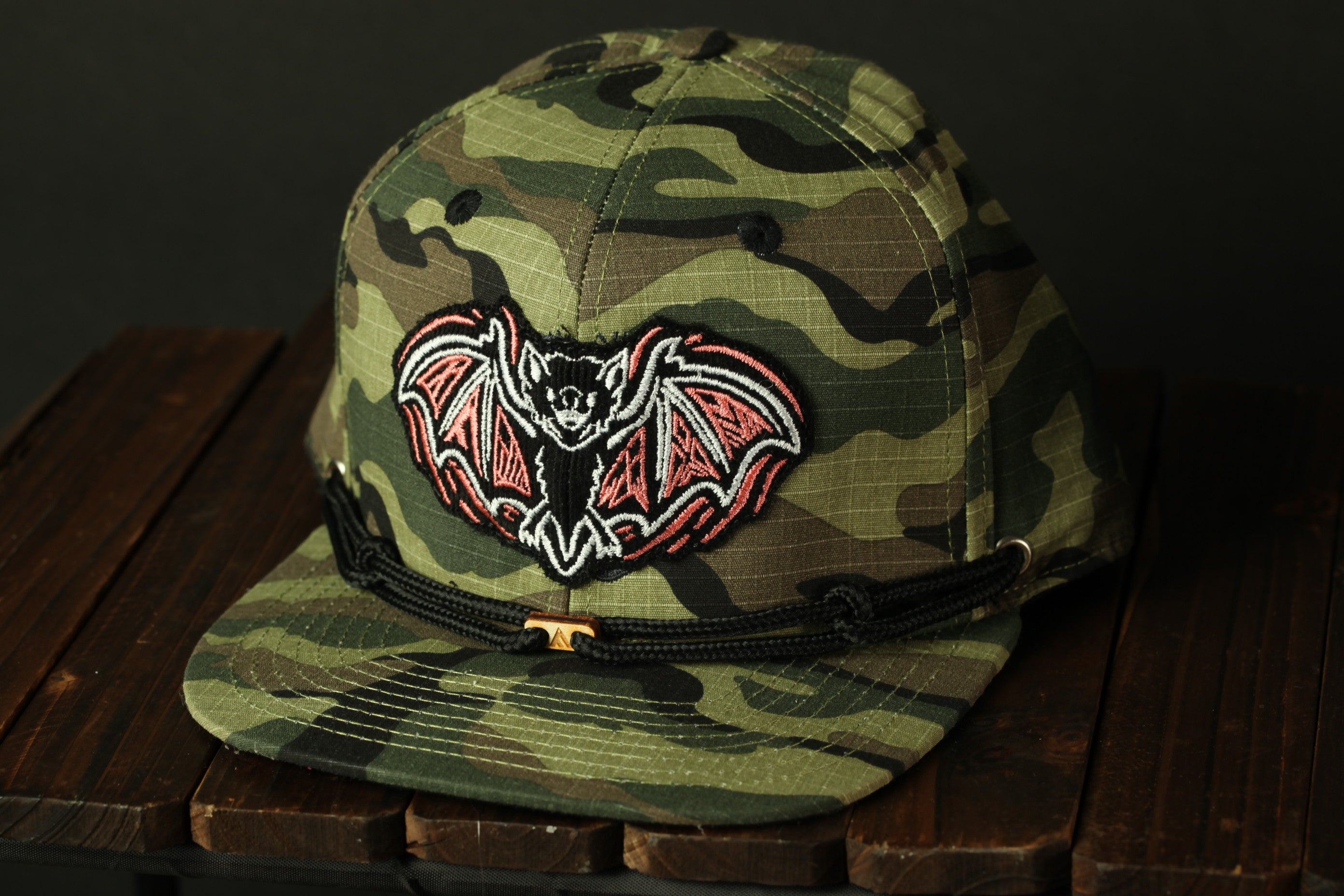 A camouflage baseball cap with a bat embroidered on it, placed on a wooden surface.