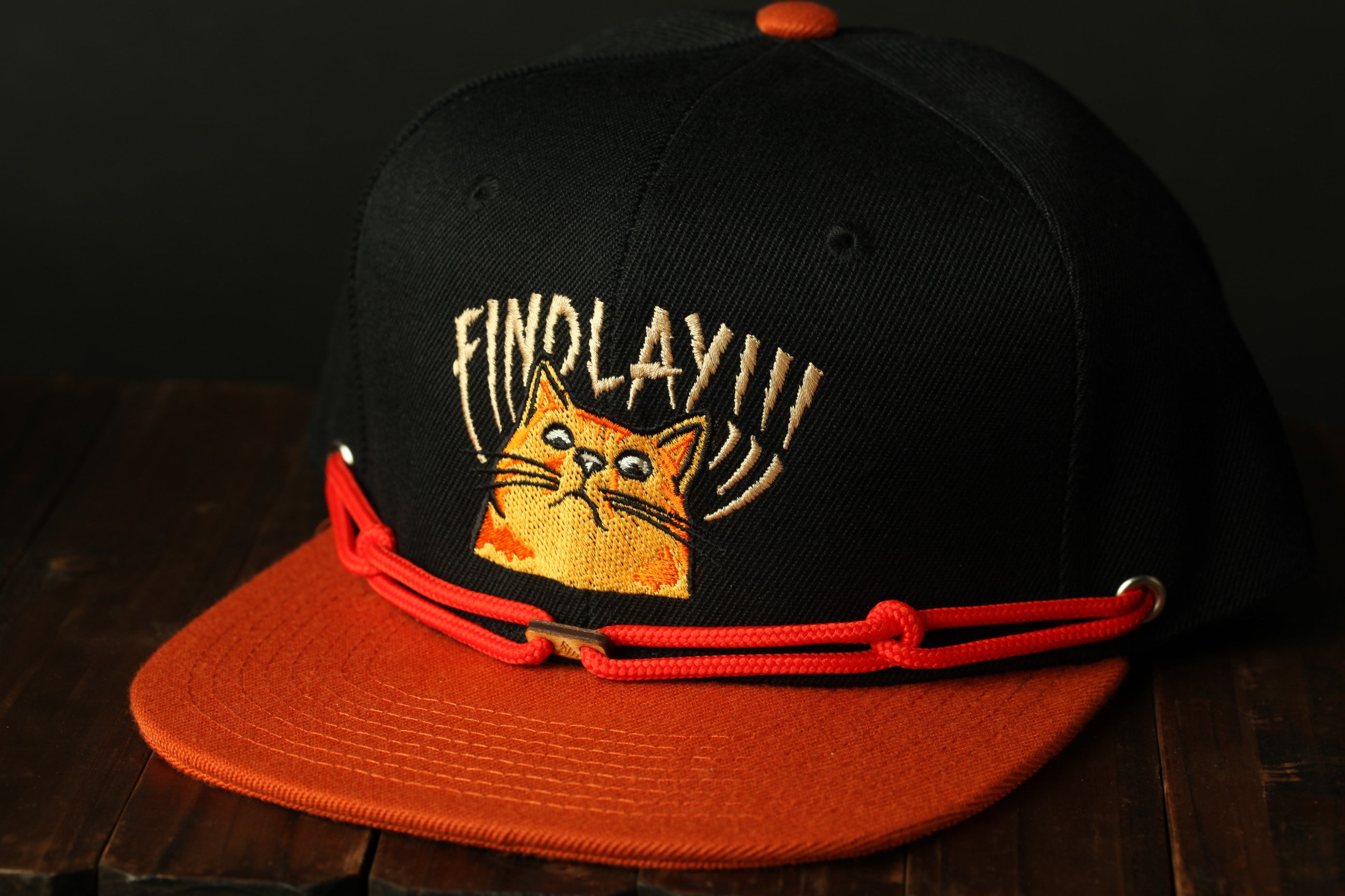 A black baseball cap with an orange brim and a cat embroidered on it, sitting on a wooden surface.