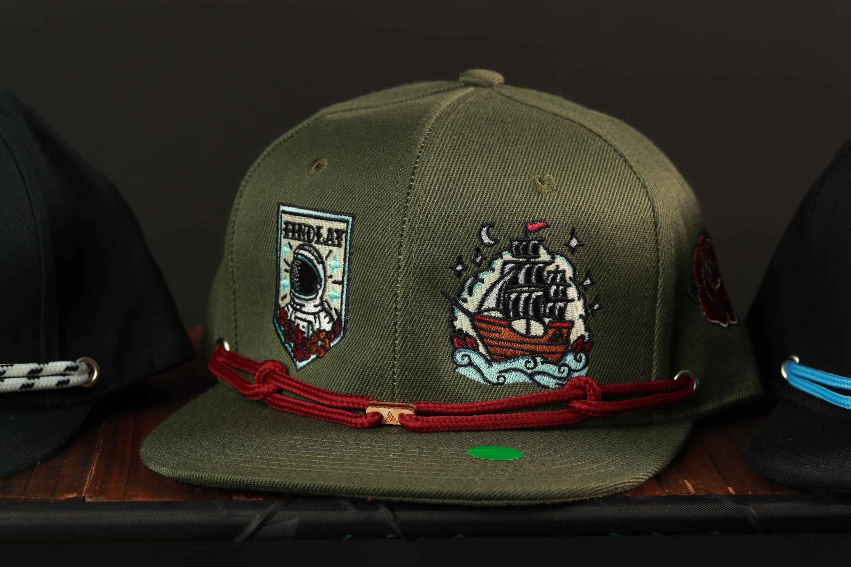 A green baseball cap with various embroidered designs, including a ship, a person, and other decorative elements, is displayed on a shelf.