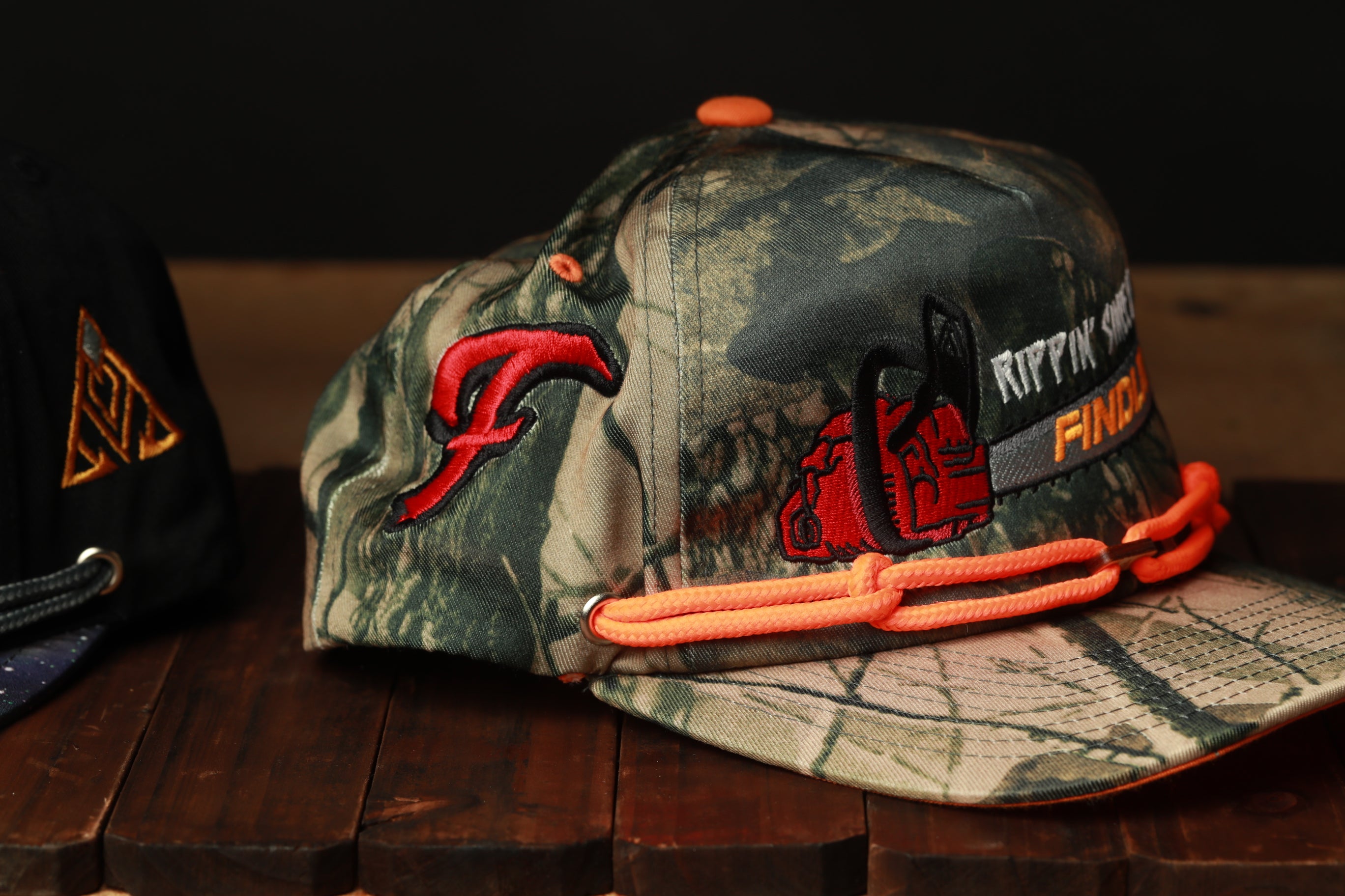 A camouflage baseball cap with a red logo and orange straps, placed on a wooden surface.