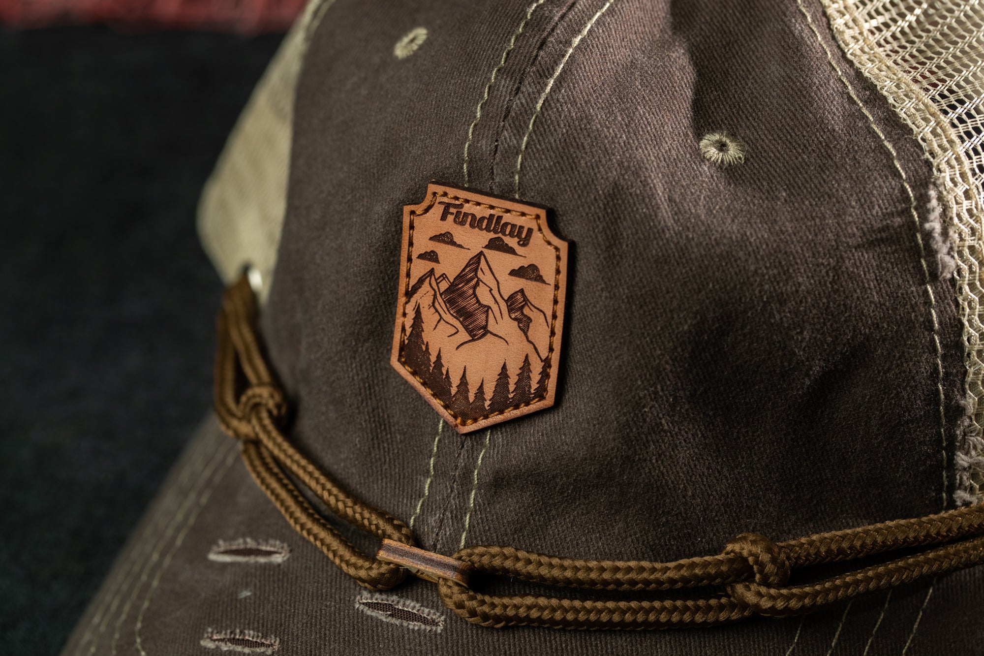 A close-up view of a brown baseball cap with a leather patch featuring a mountain landscape and the word "Findlay" printed on it.