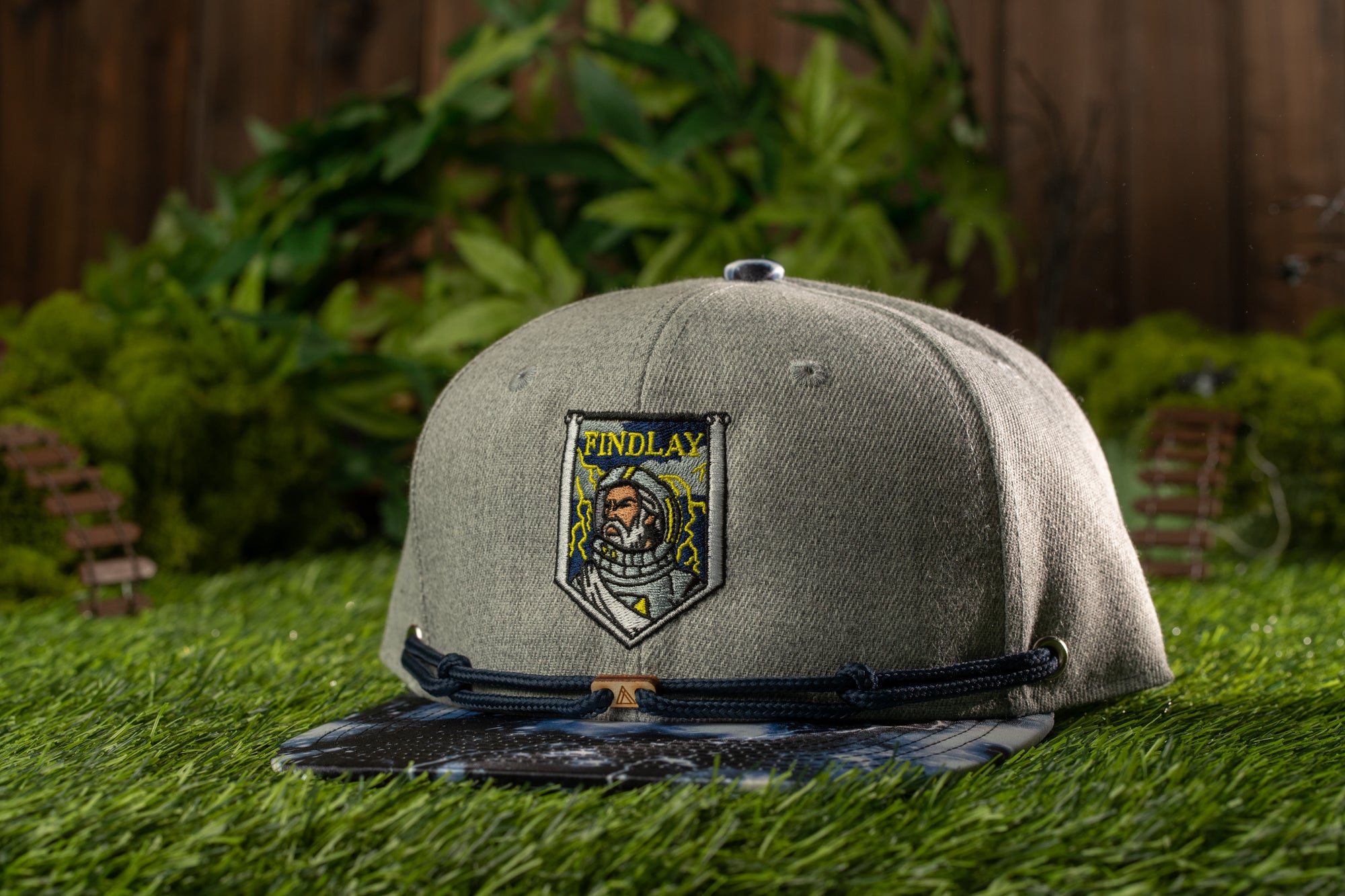 A gray baseball cap with a black band and a patch featuring a cartoon character is placed on a grassy surface.