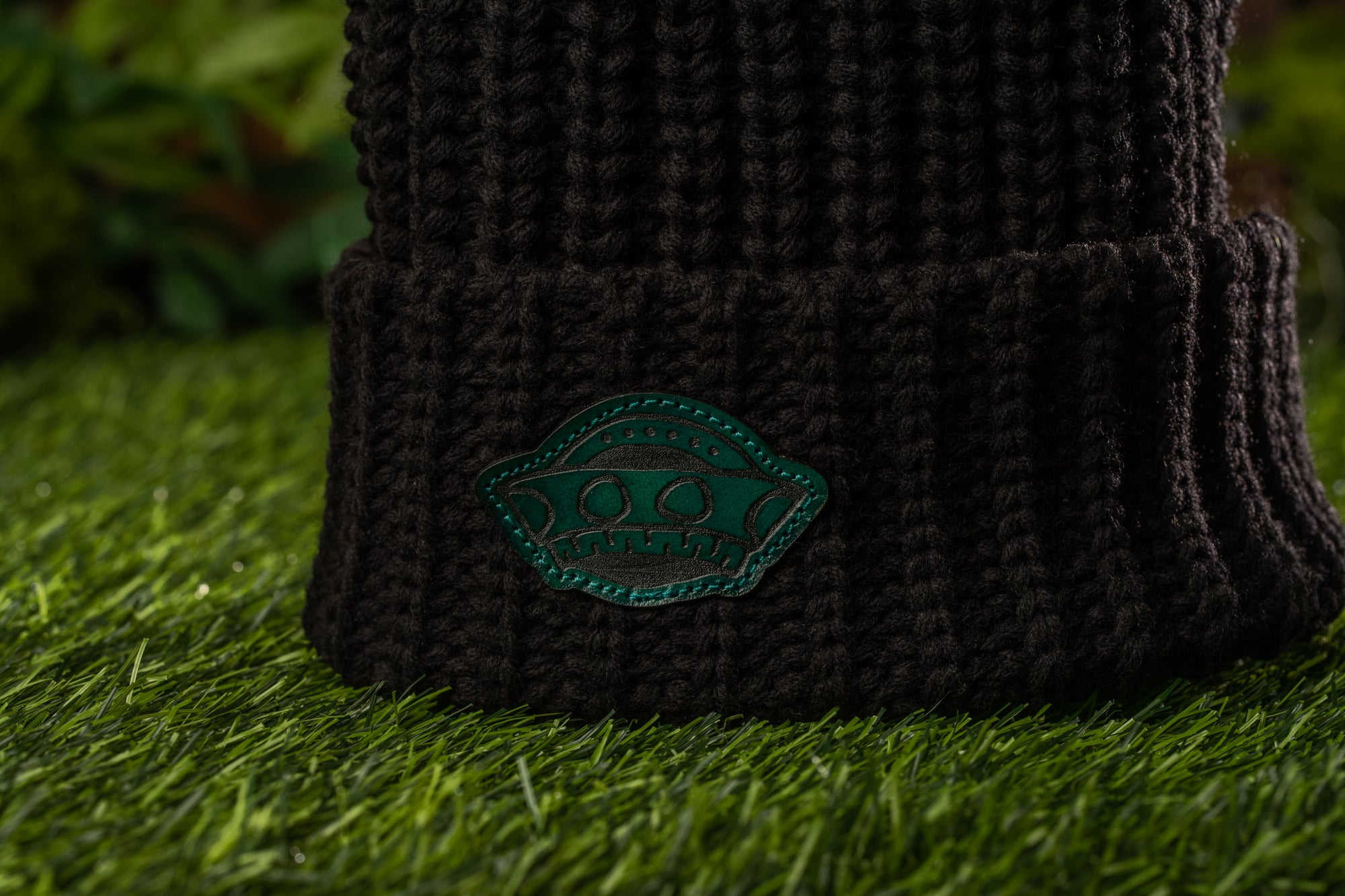 A close-up view of a dark knit beanie with a green patch on the front, placed on a grassy surface.
