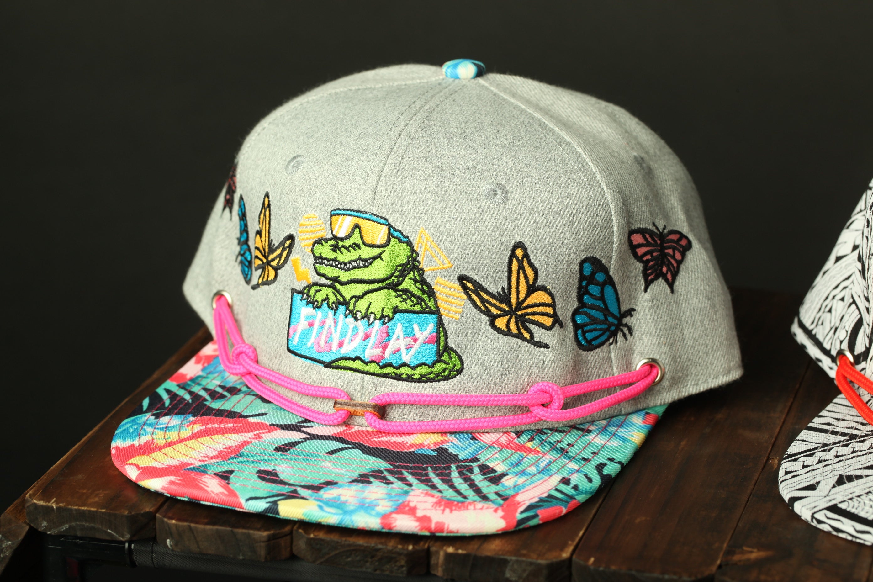 A gray baseball cap with a colorful floral pattern and a cartoon character on the front, sitting on a wooden surface.