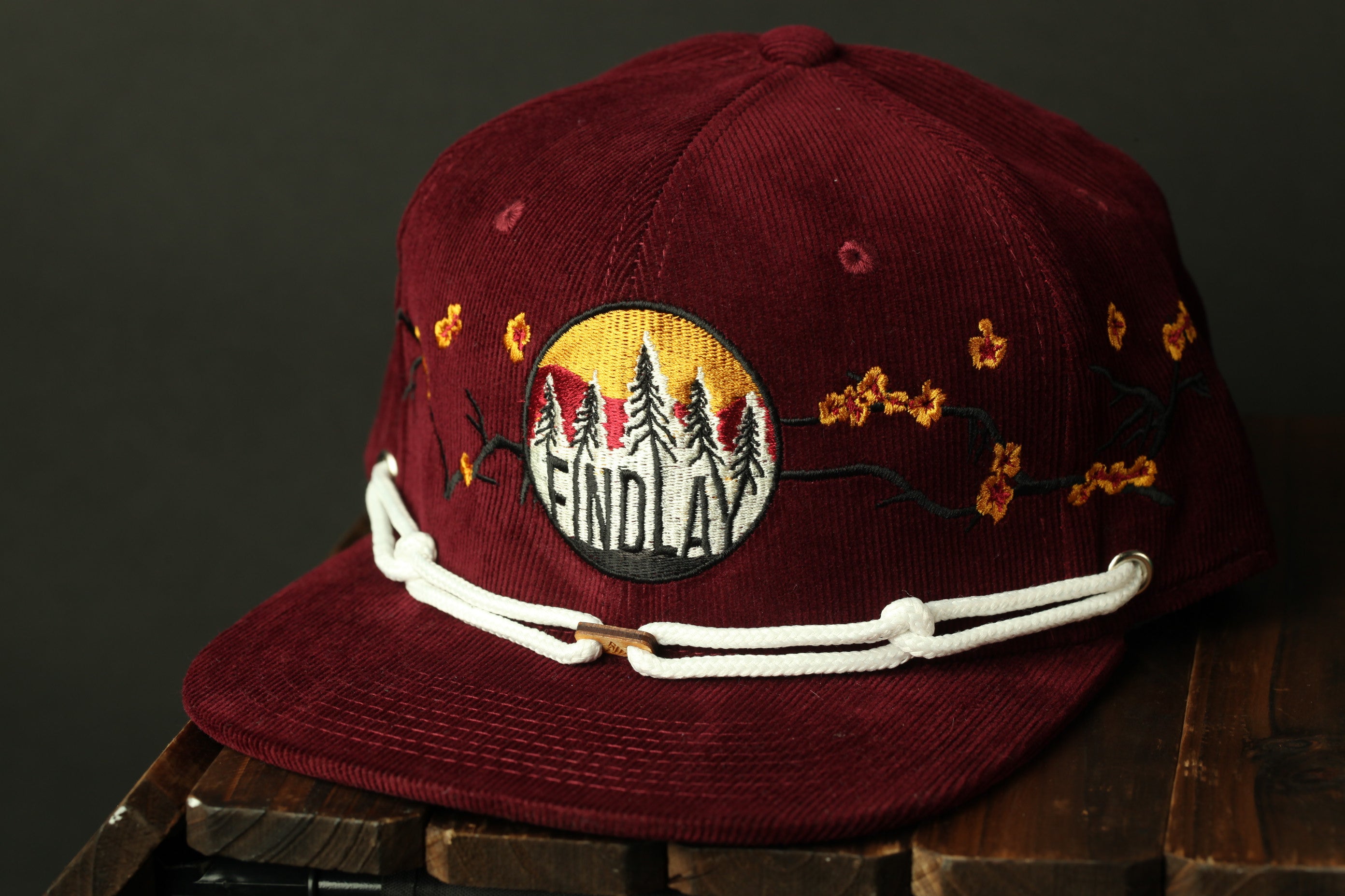 A burgundy baseball cap with a white lace trim and a circular patch on the front, featuring a design with trees and flowers.