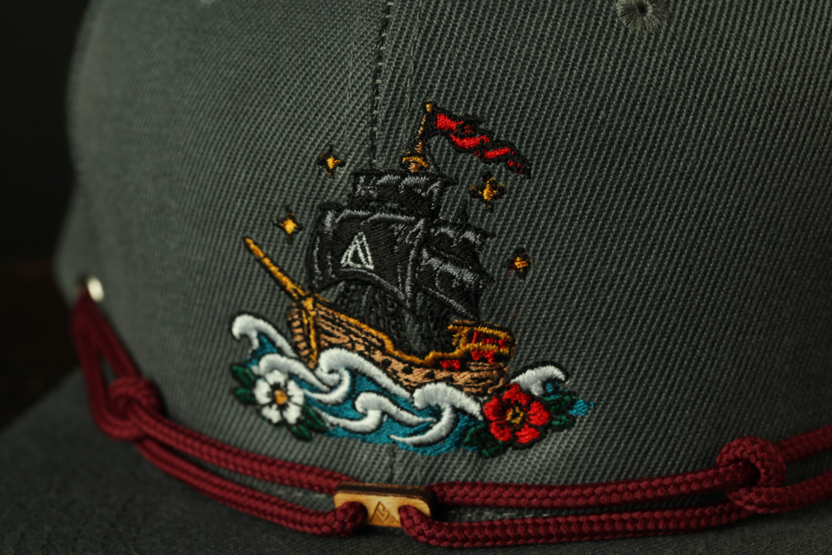a close-up view of a gray baseball cap with a detailed embroidered design on the front. The design depicts a ship sailing on a body of water, surrounded by waves and flowers.