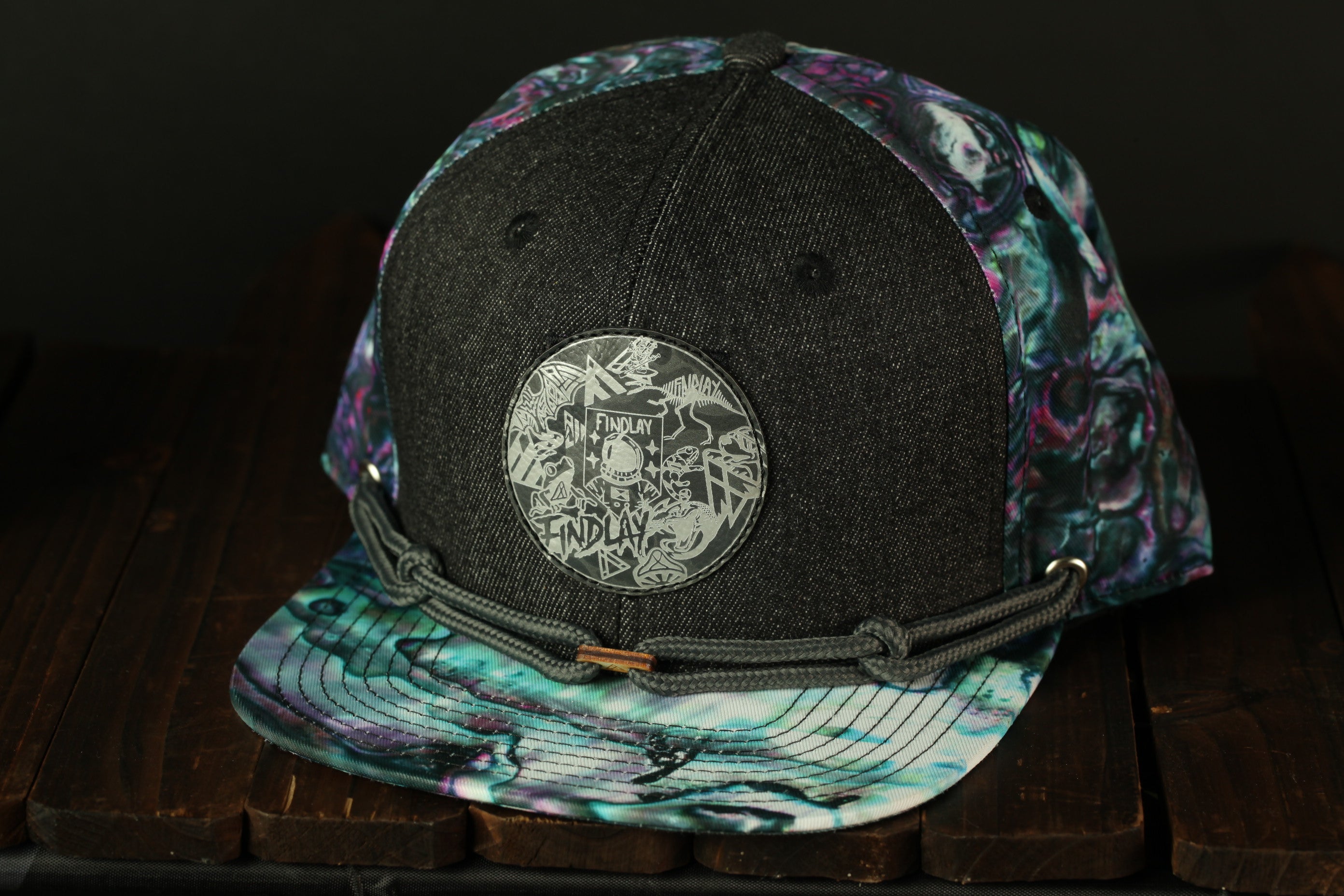 A black baseball cap with a colorful, abstract pattern on the front and a circular patch on the front, sitting on a wooden surface.