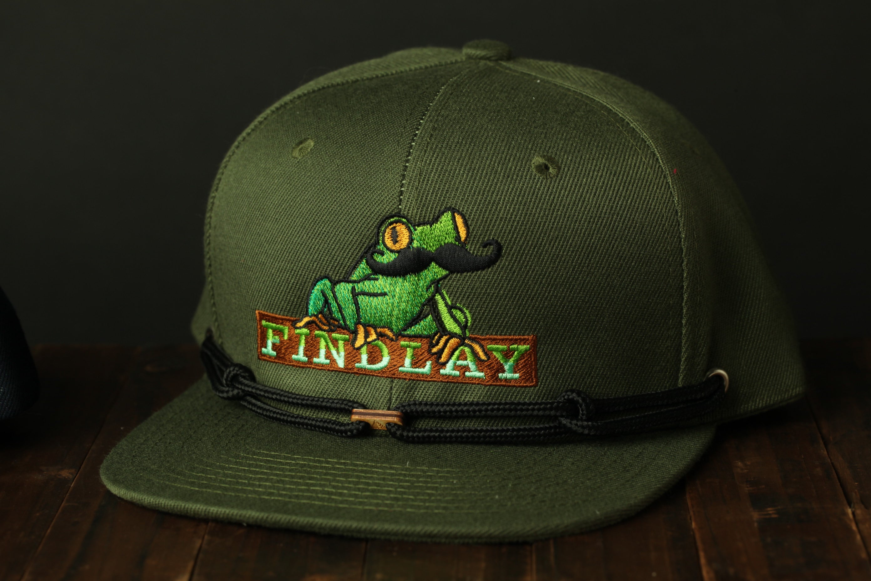 A green baseball cap with a frog embroidered on it, featuring a mustache and the word "FINDLAY" on the front.