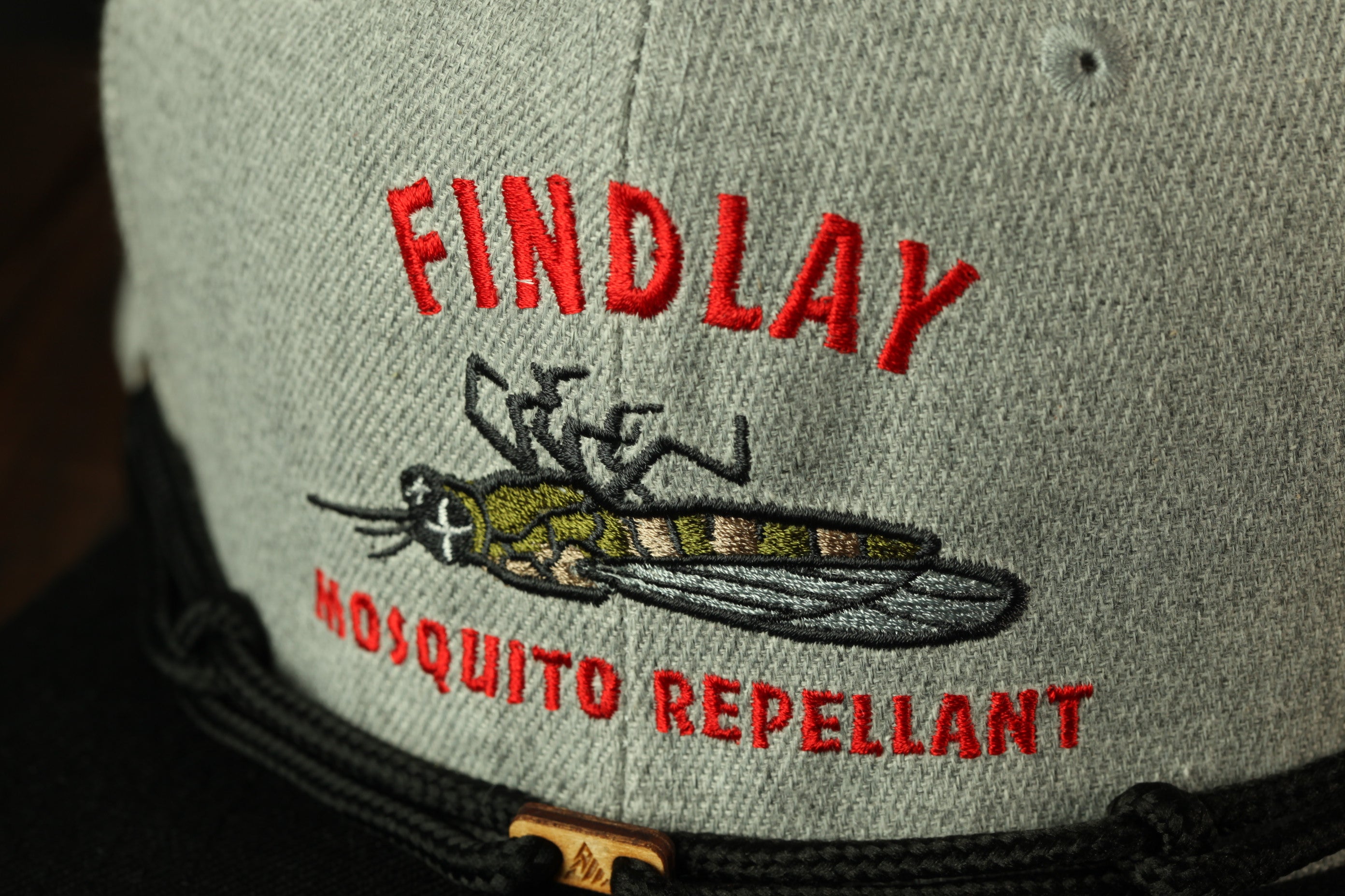 a gray baseball cap with a red and black embroidered design on the front. The design includes a close-up of a mosquito and the text "FINDLAY" and "MOSQUITO REpellent" printed on it.