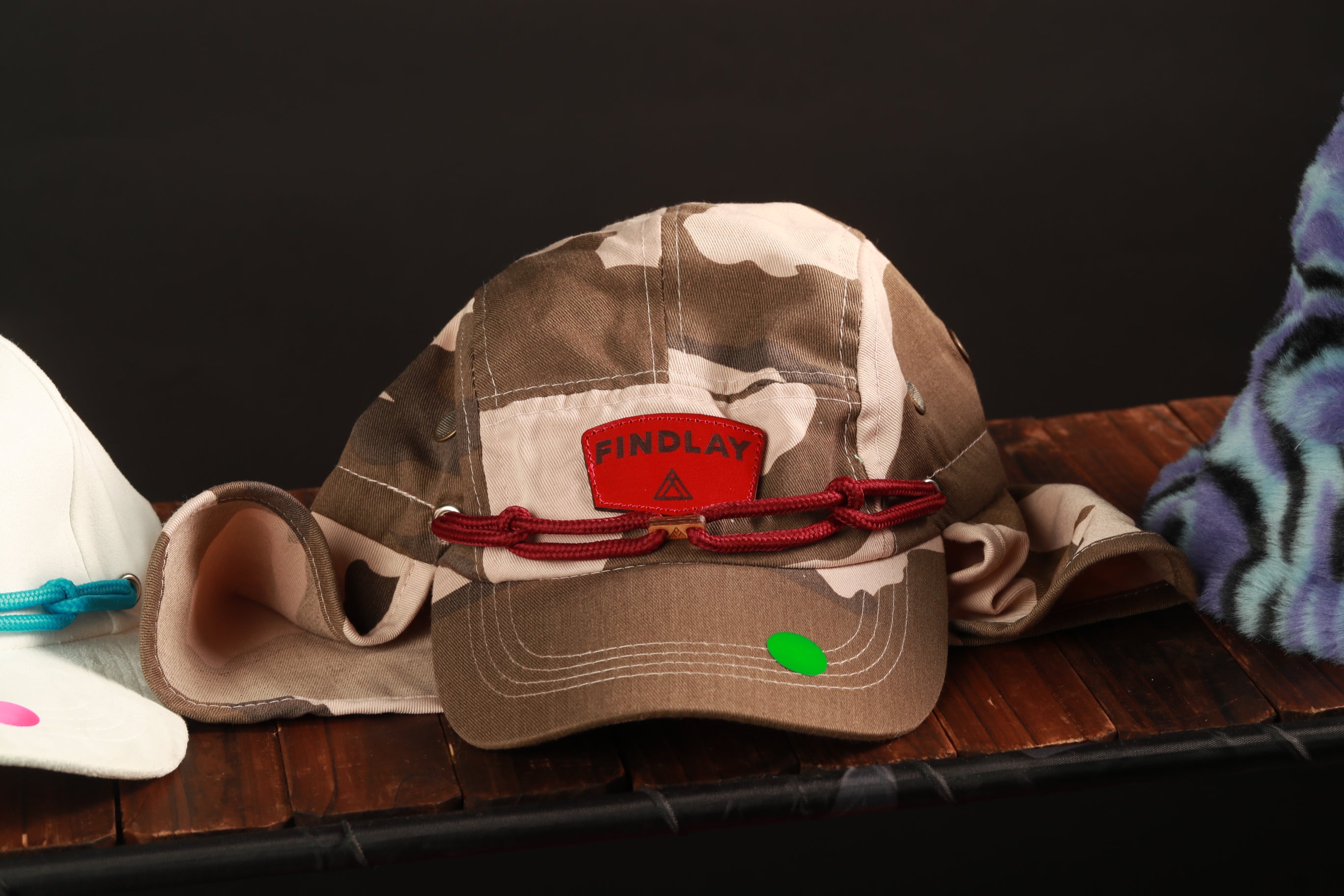 A camouflage baseball cap with a red "Findlay" patch and a green tag is displayed on a wooden surface.