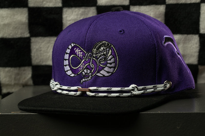 A purple baseball cap with a snake design on it, sitting on a black surface against a checkered background.