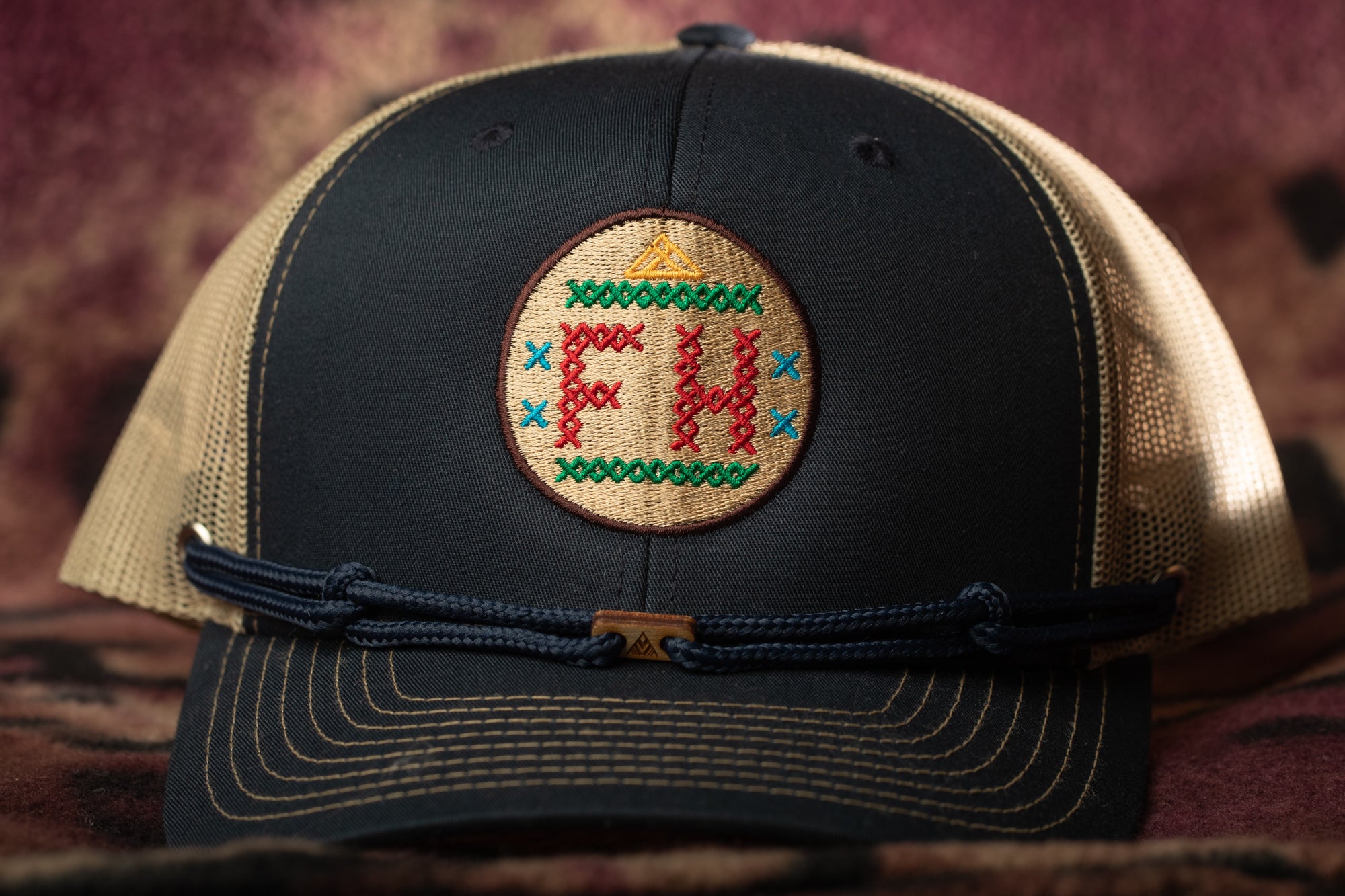 A black baseball cap with a beige mesh panel and a circular embroidered patch on the front.