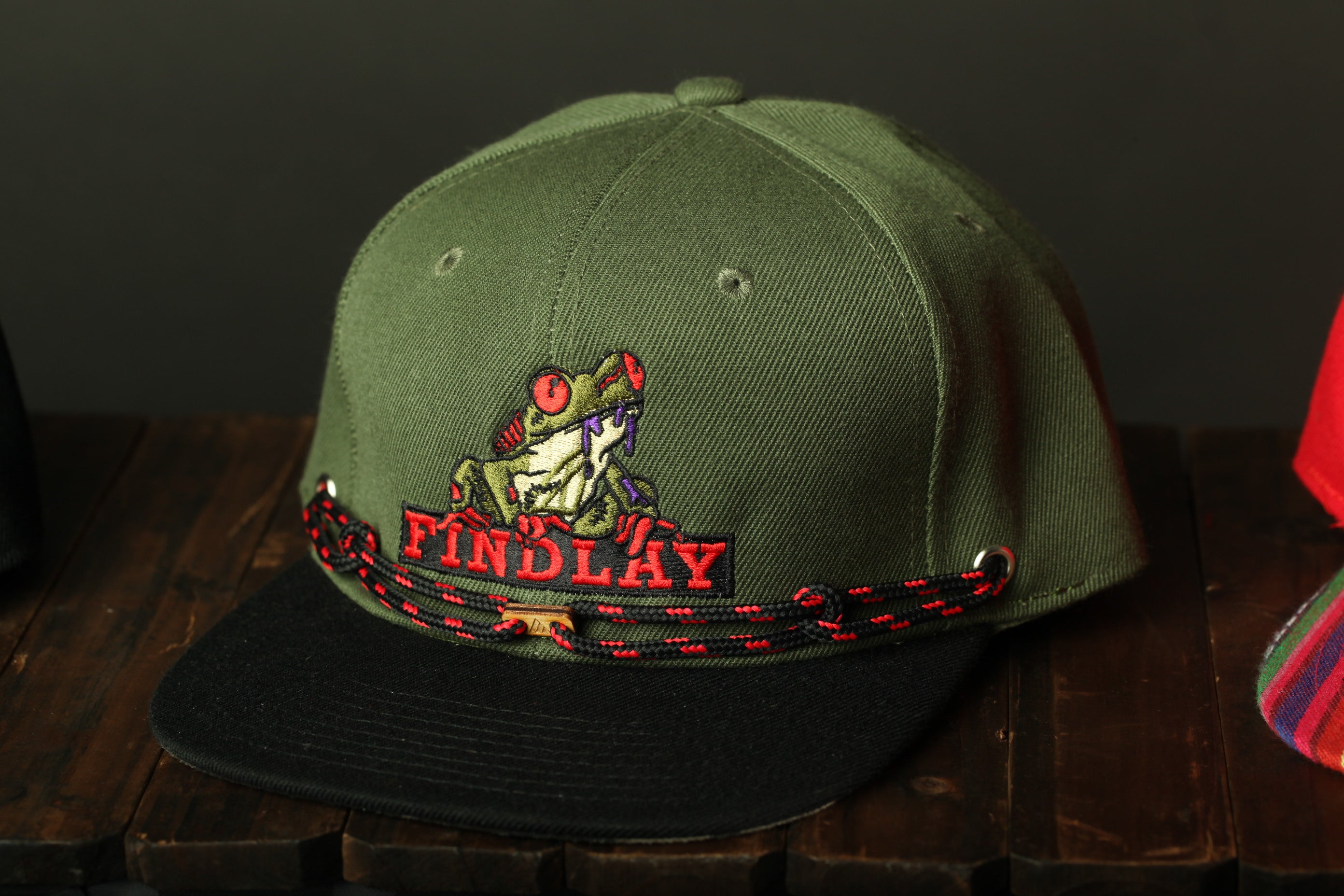 A green baseball cap with a red and black braided strap and a cartoon frog logo on it, placed on a wooden surface.