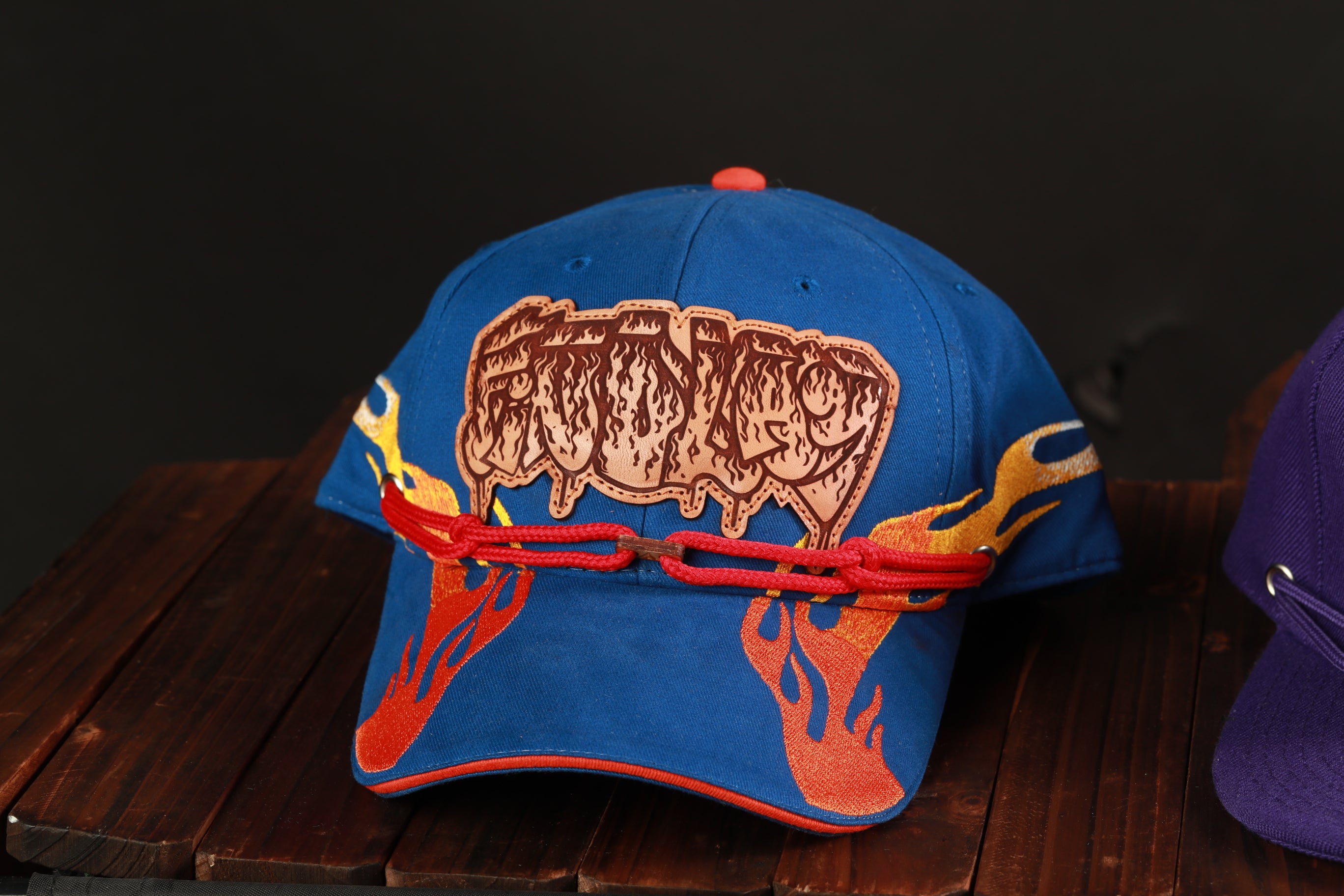 A blue baseball cap with a flame design and a red chain attached to it, placed on a wooden surface.