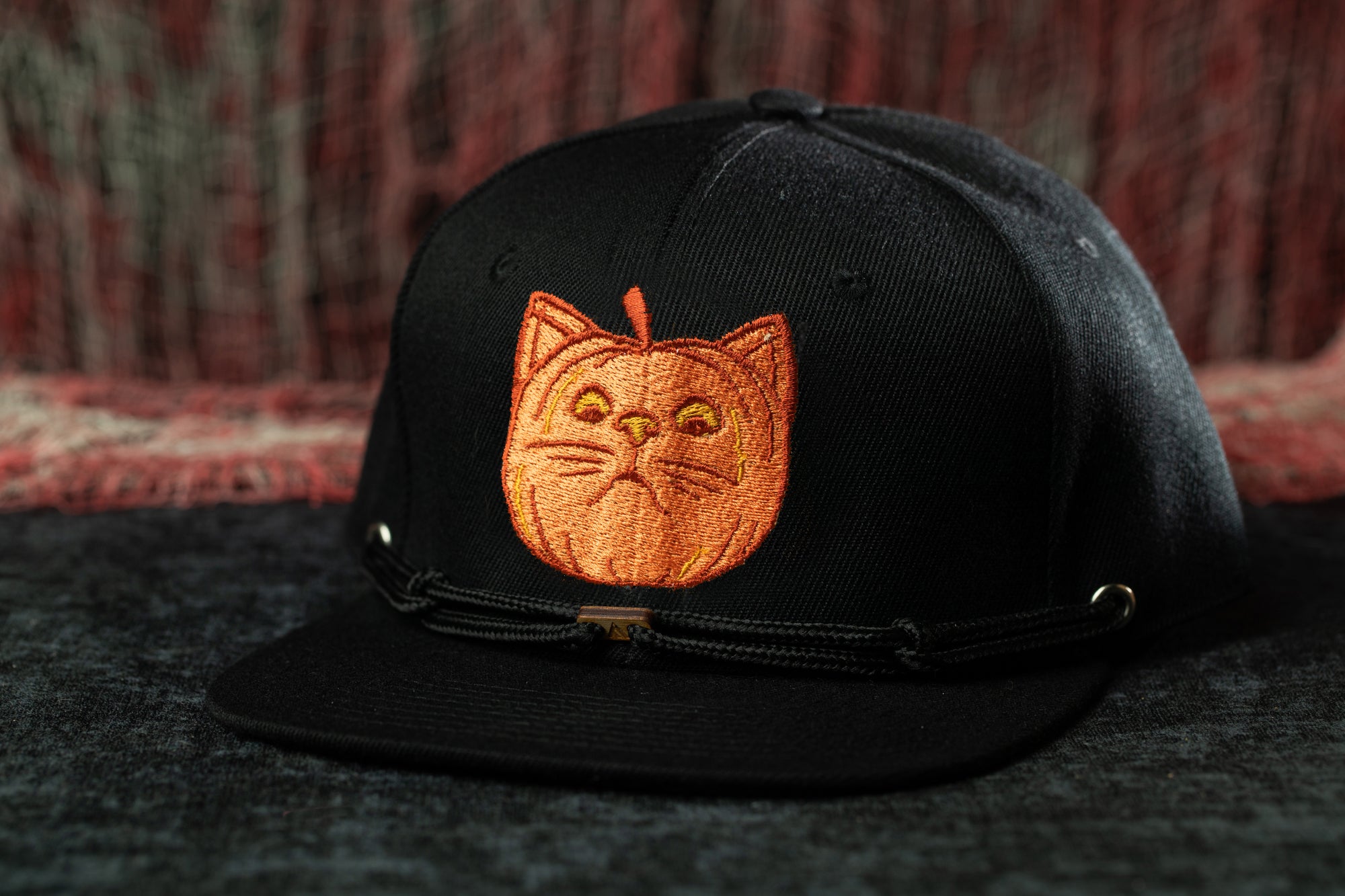 A black baseball cap with an orange cat embroidered on the front, sitting on a dark surface.