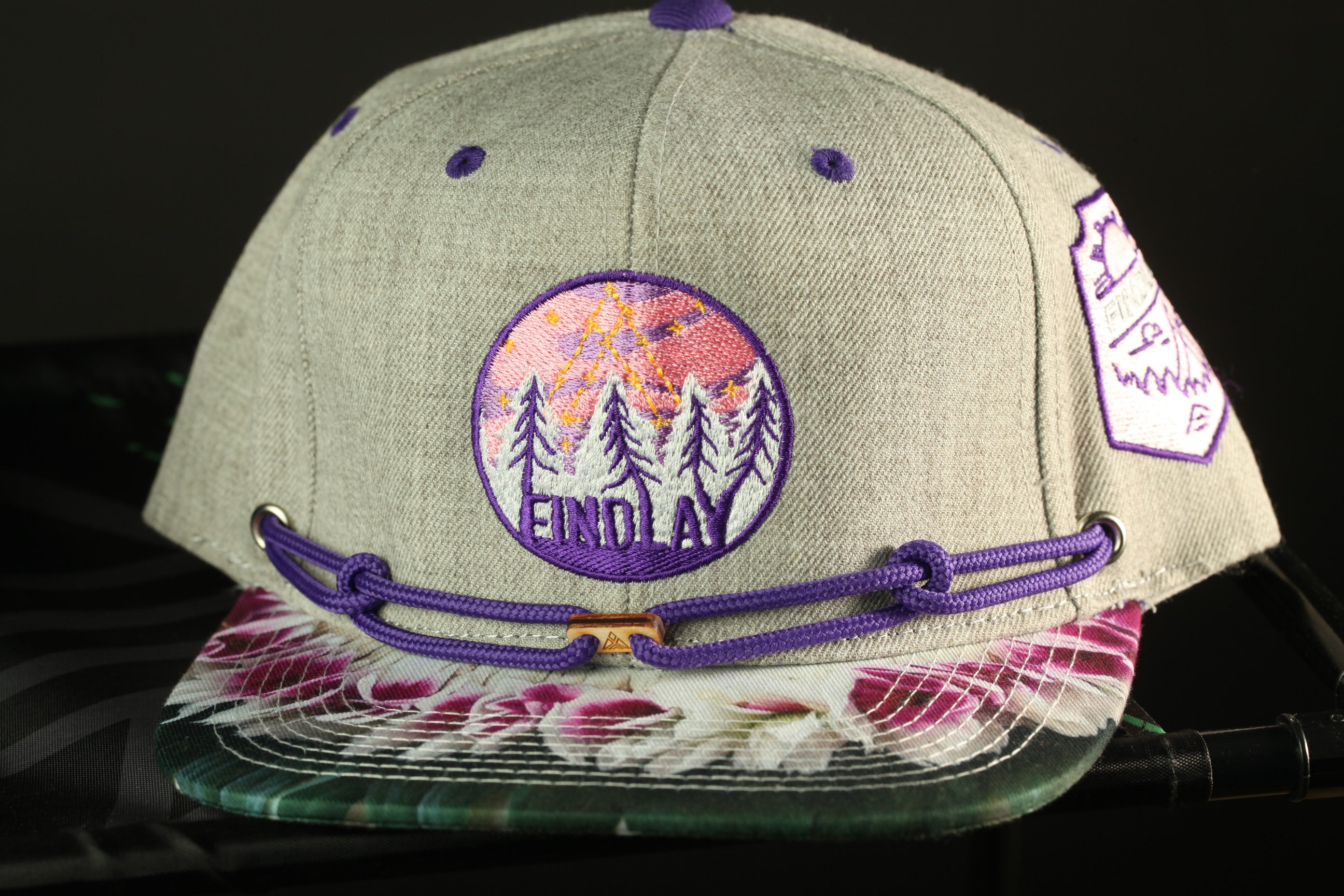 A gray baseball cap with purple and white floral patterns, a logo, and a purple string.