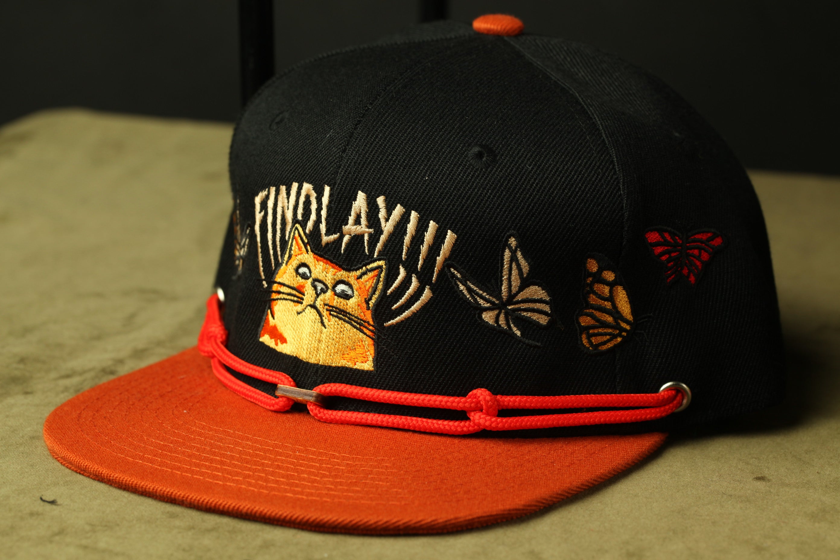 A black baseball cap with an orange brim and a cat embroidered on it, along with some butterflies and a red bow.