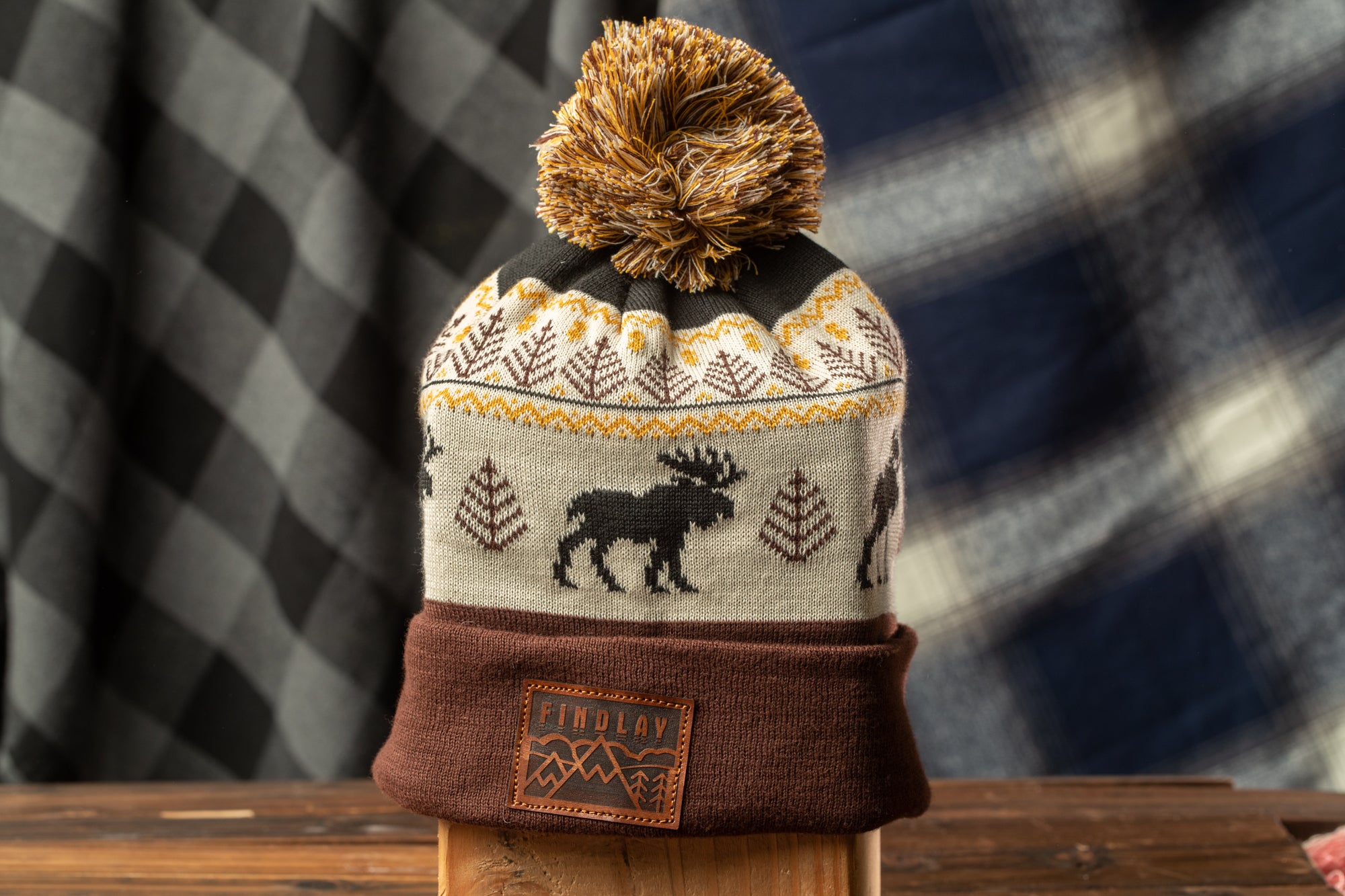 A brown and white knit beanie with a pom pom on top, featuring a moose design, is displayed on a wooden surface against a background of a checkered fabric.