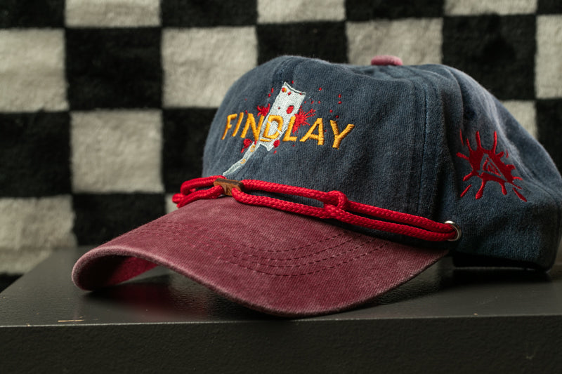 A maroon baseball cap with a red string and the word "FINOLAY" printed on it, placed on a black surface against a checkered background.