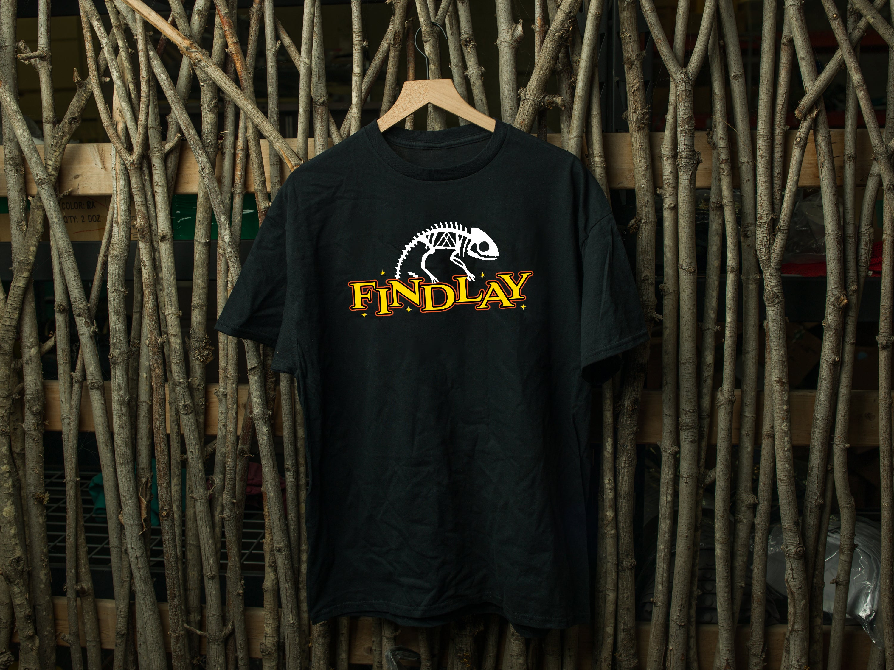 A black t-shirt with a graphic design and text printed on it, hanging on a wooden hanger against a background of wooden sticks or branches.