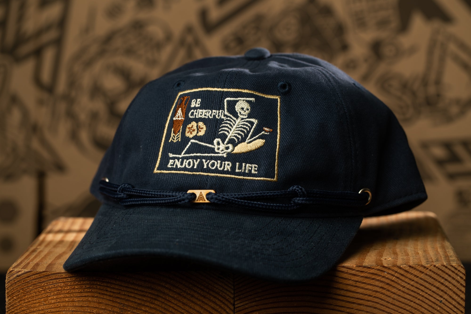 A navy blue baseball cap with a skull and crossbones design and the text "Be Cheerful, Enjoy Your Life" printed on it.