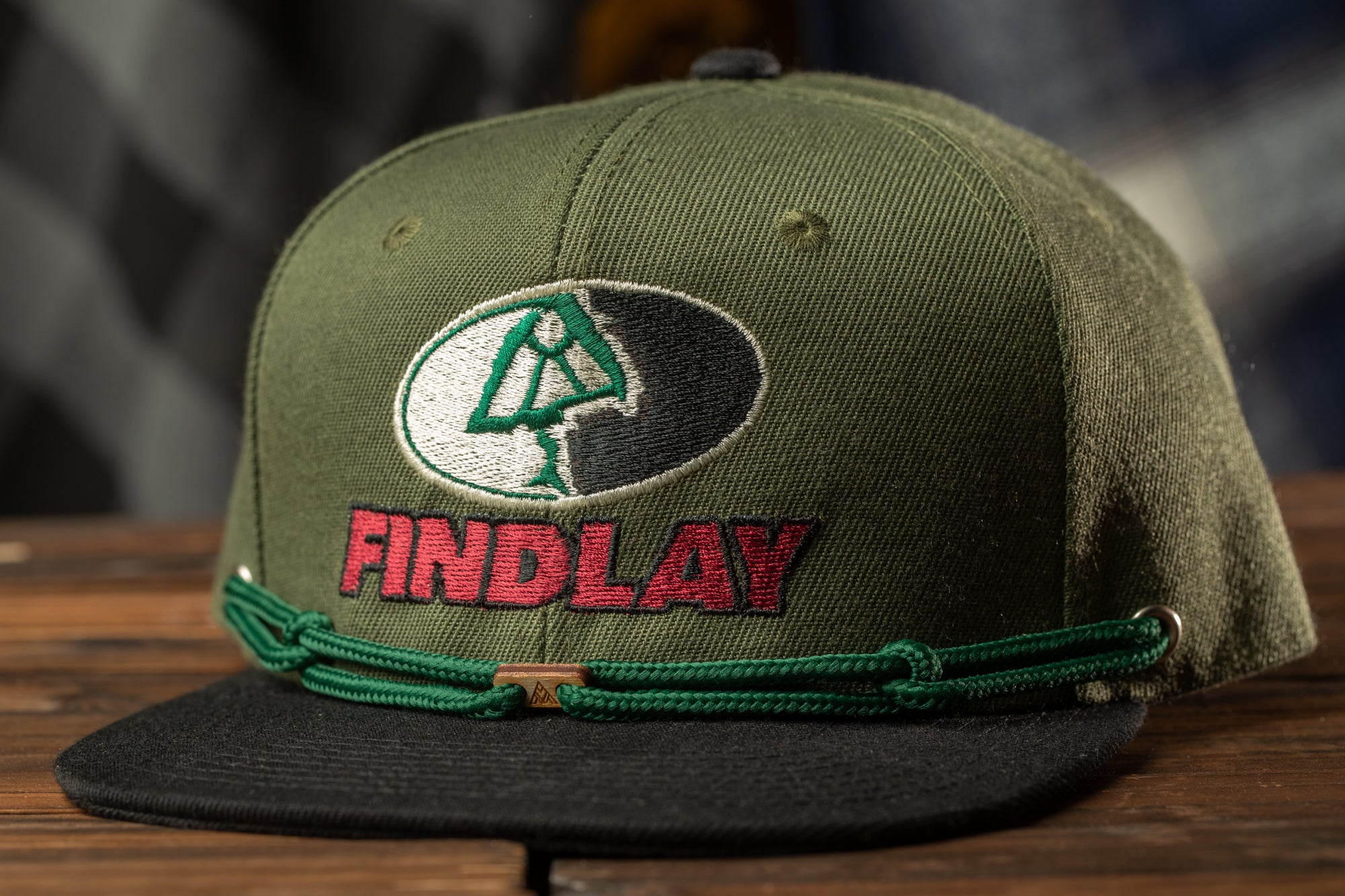 A green baseball cap with a logo and the word "FINDLAY" printed on it, sitting on a wooden surface.