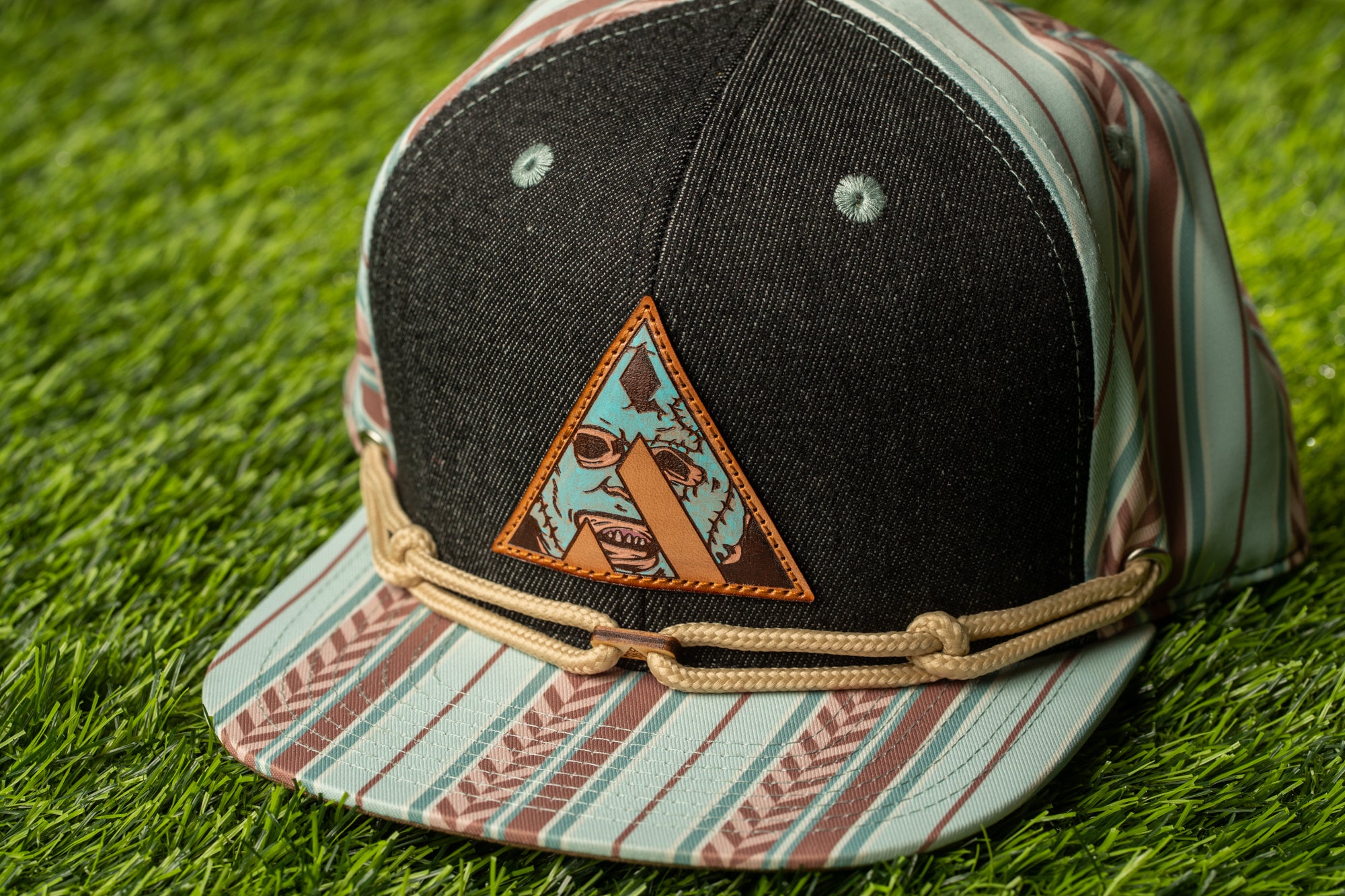 A black baseball cap with a colorful, cartoonish design on the front, featuring a triangle with a face and a rope around the brim.