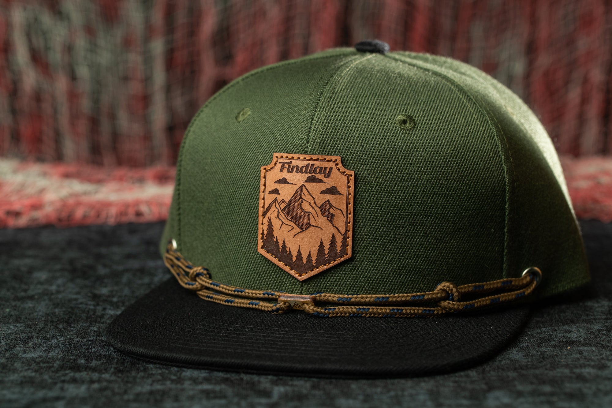 A green baseball cap with a leather patch on the front, featuring a mountain landscape design.