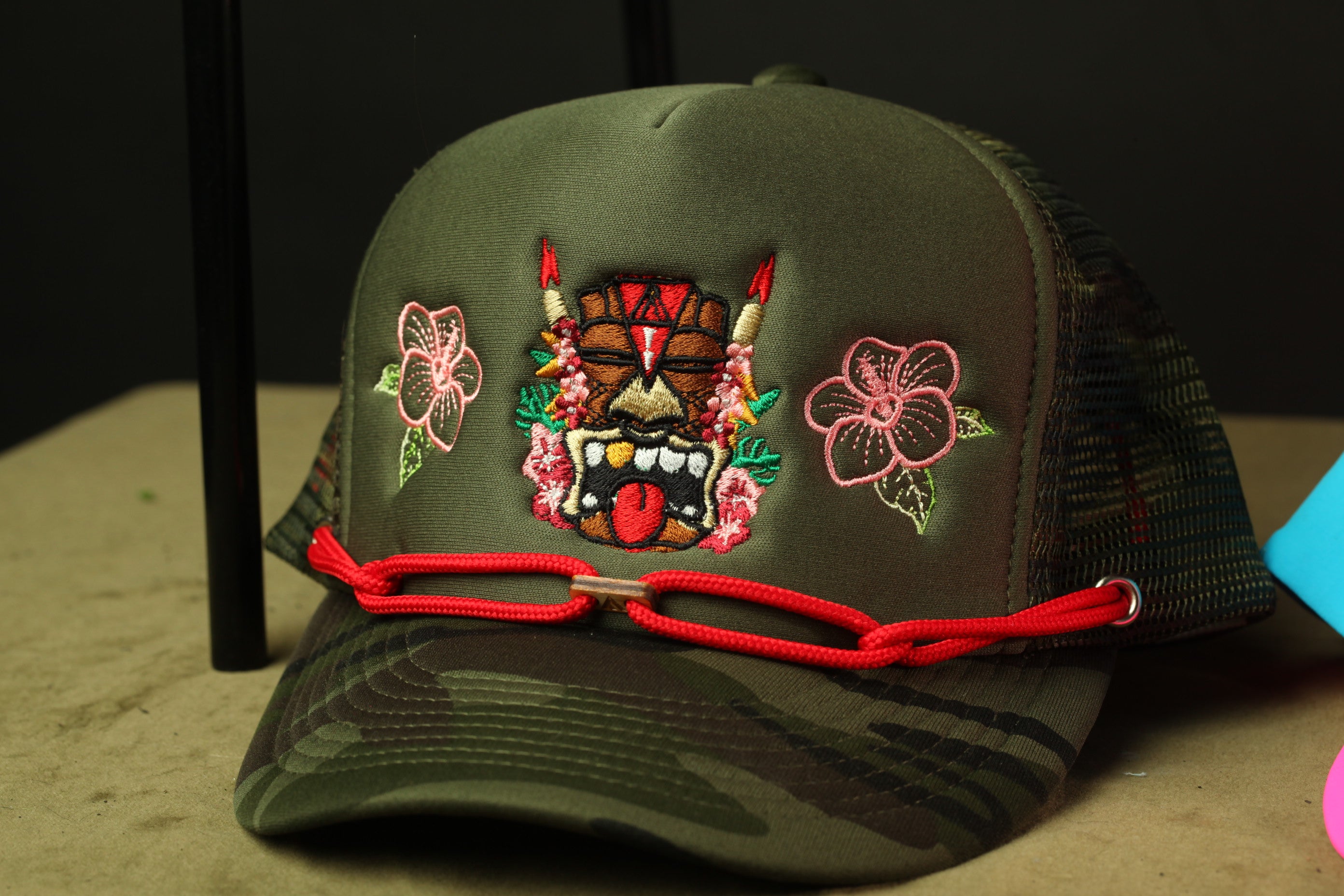 A green baseball cap with a colorful tiki mask design and flowers on the front.