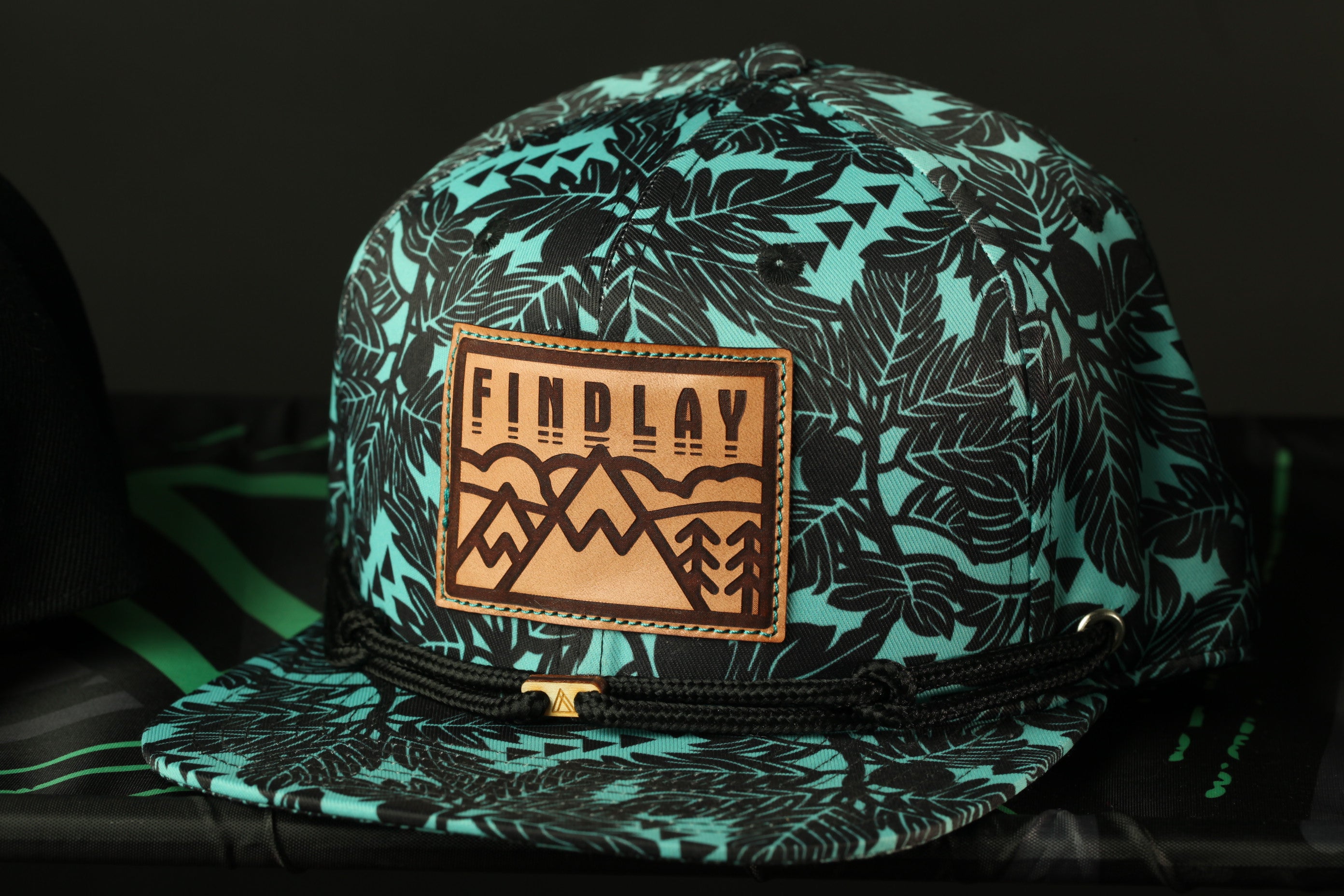 A turquoise baseball cap with a leafy pattern and a leather patch that says "FINDLAY" on it.