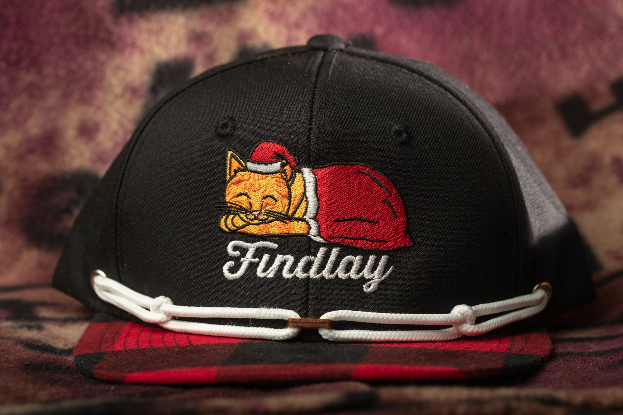 A black baseball cap with a red and white plaid pattern and a cat embroidered on it.