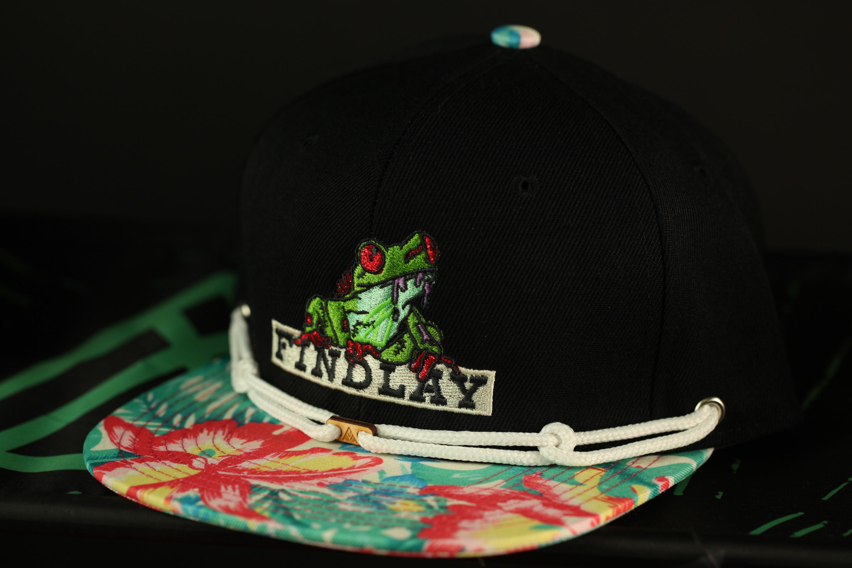 A black baseball cap with a colorful floral pattern and a frog embroidered on it, sitting on a surface with a green and red patterned fabric underneath.