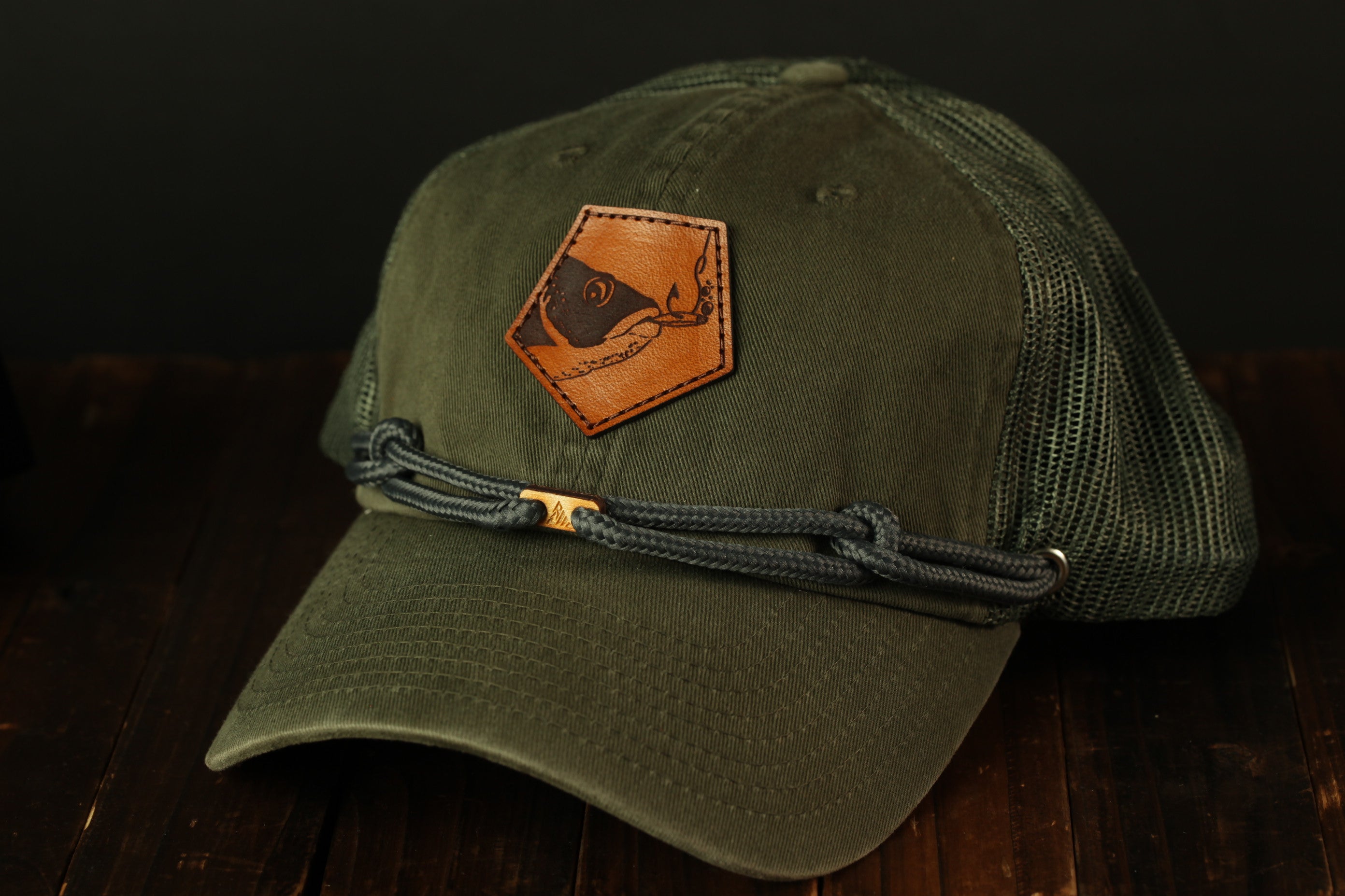 A green baseball cap with a leather patch featuring a fish design, resting on a wooden surface.