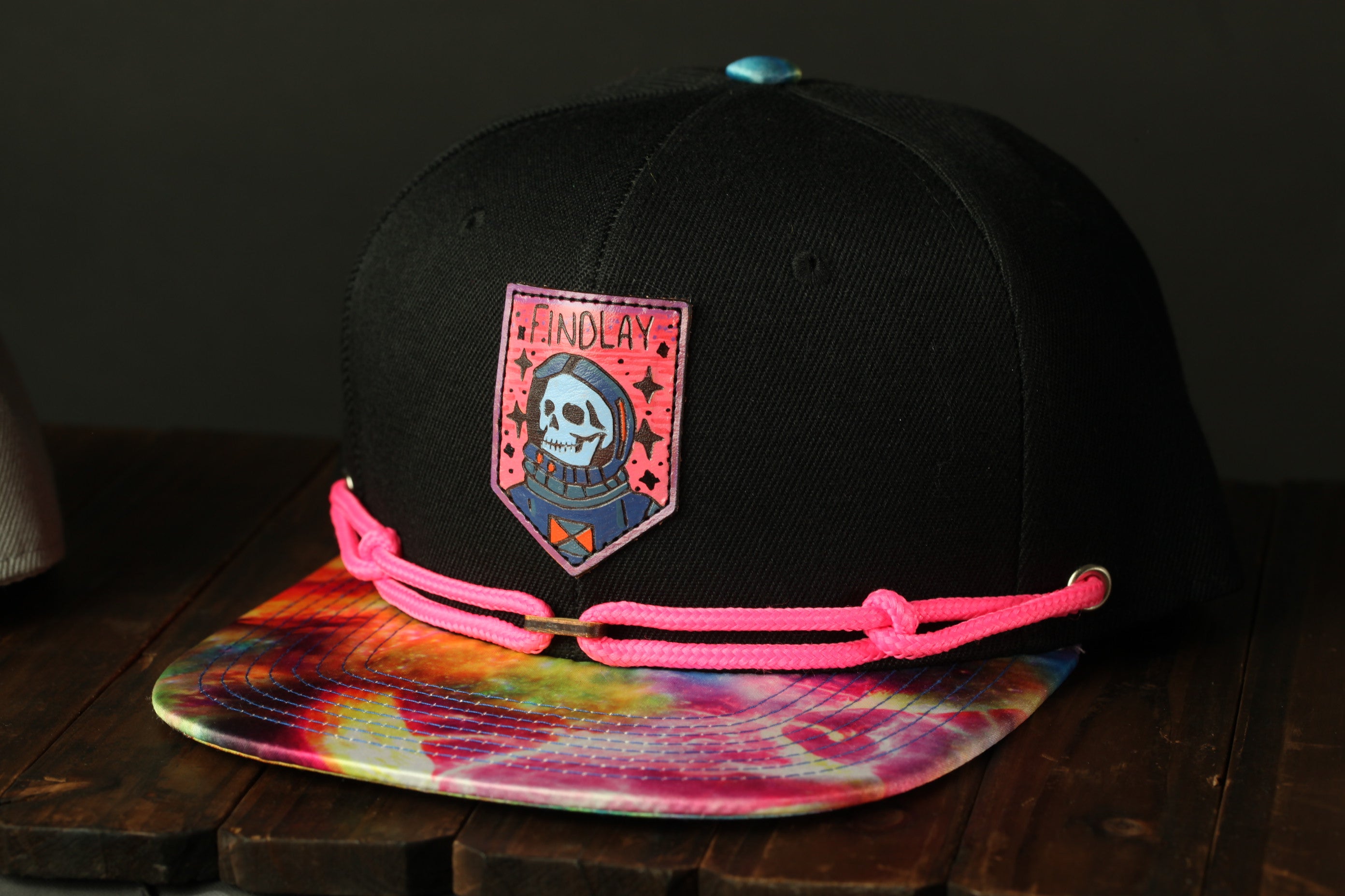 A black baseball cap with a colorful tie-dye pattern and a skull patch on the front, sitting on a wooden surface.